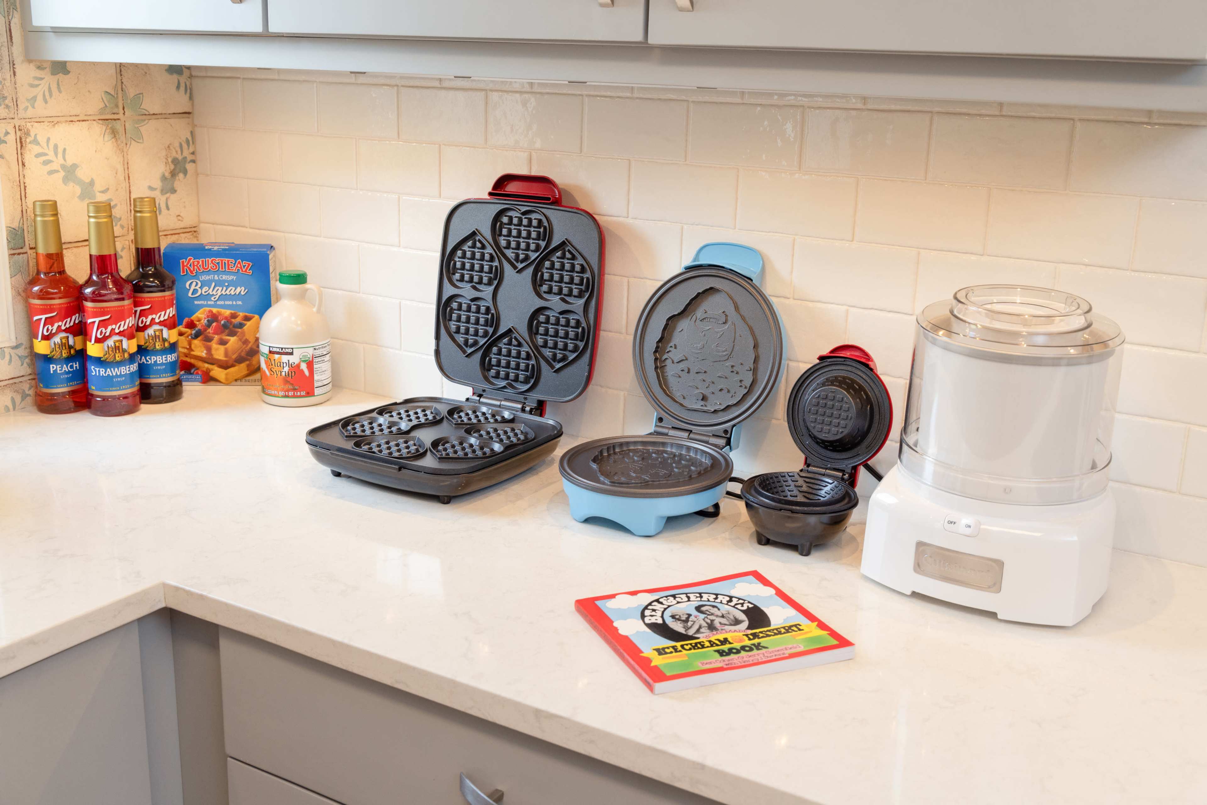 A kitchen countertop displays a waffle maker, an ice cream maker, and various bottles of syrup alongside a box of waffle mix.
