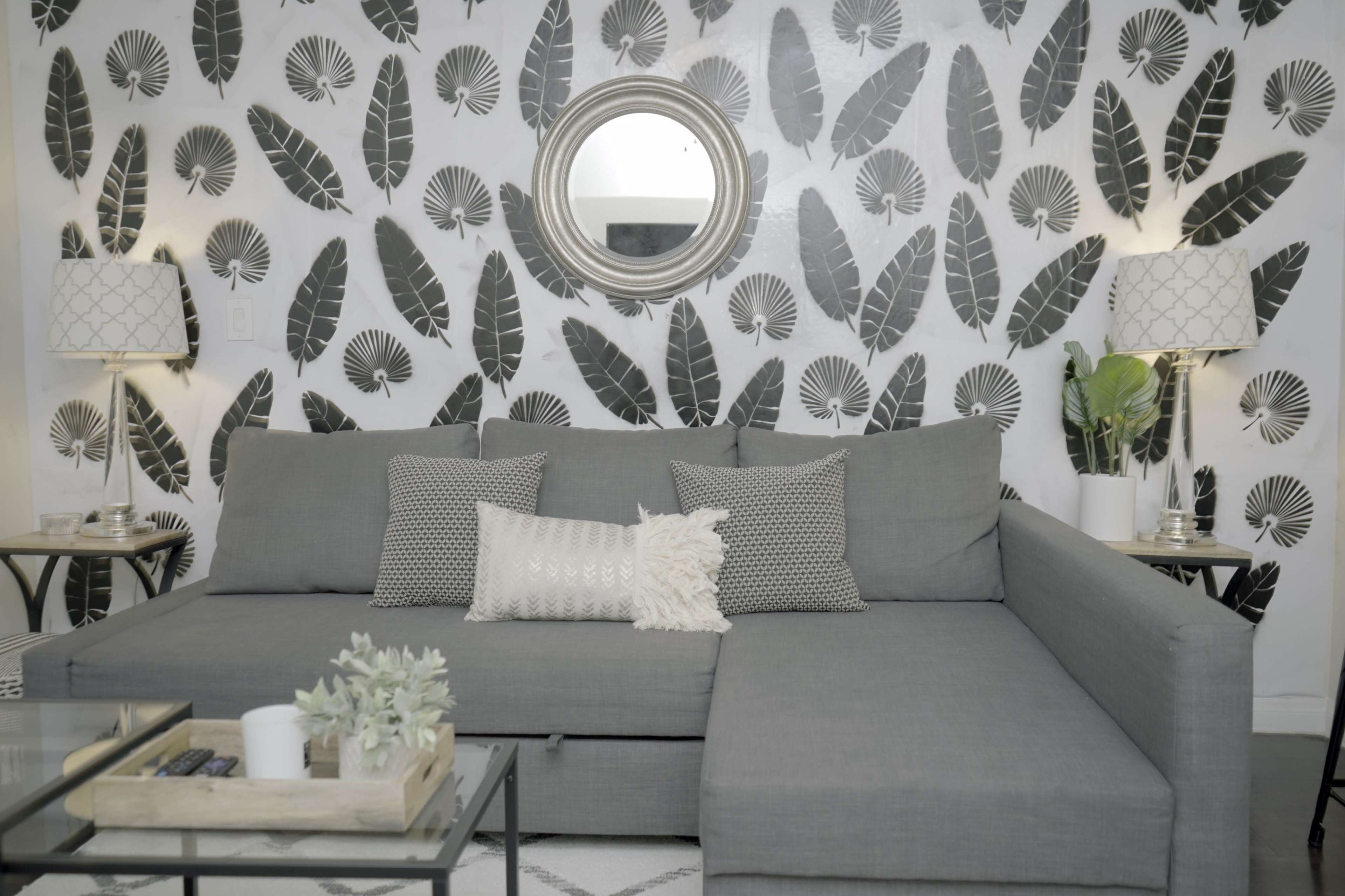 A gray sectional sofa with decorative pillows sits in front of a wall adorned with leaf-patterned wallpaper, accompanied by a round mirror and side tables with lamps.