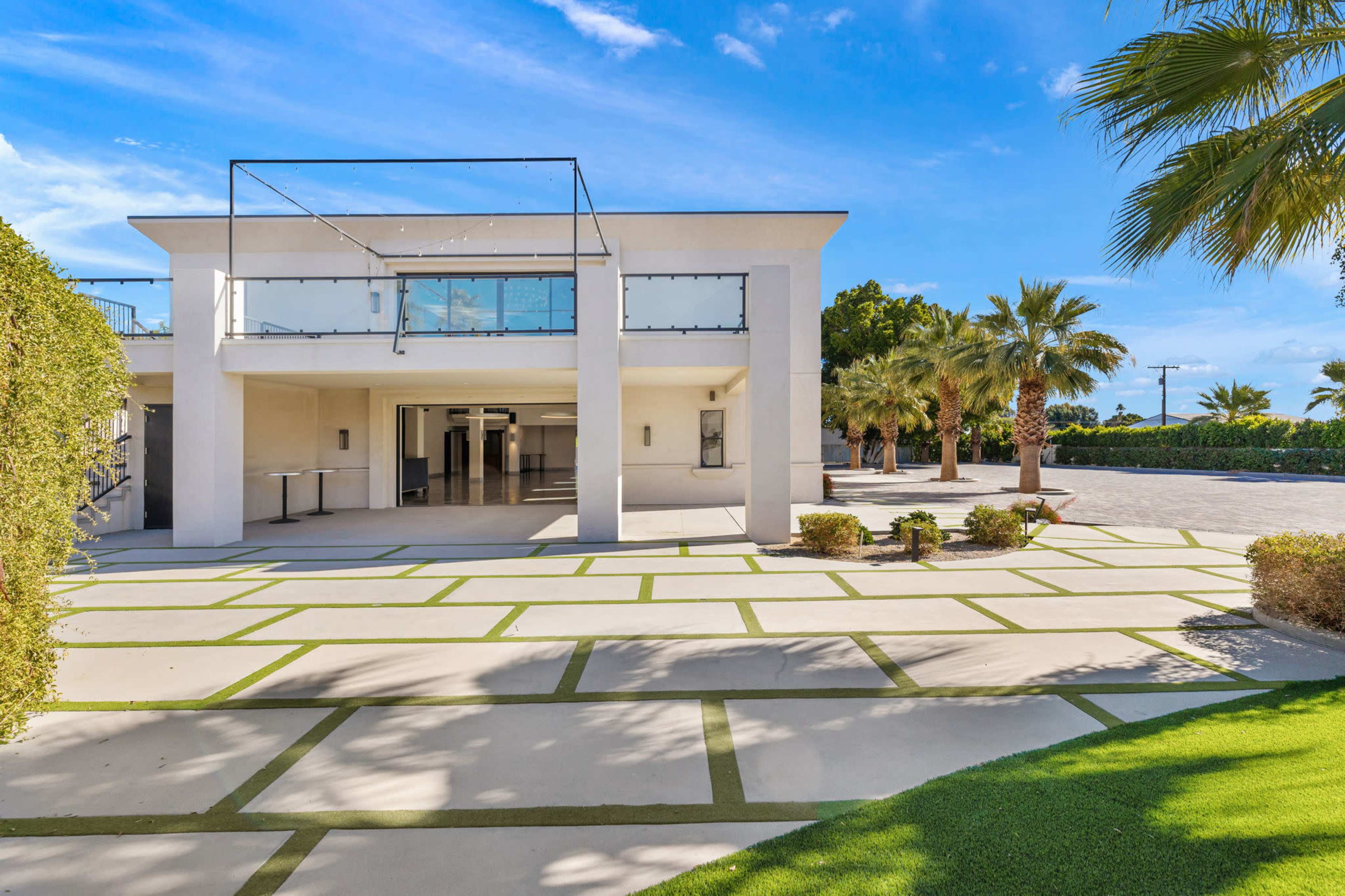 A modern two-story building with a flat roof is surrounded by palm trees and features a paved outdoor area with green accents.
