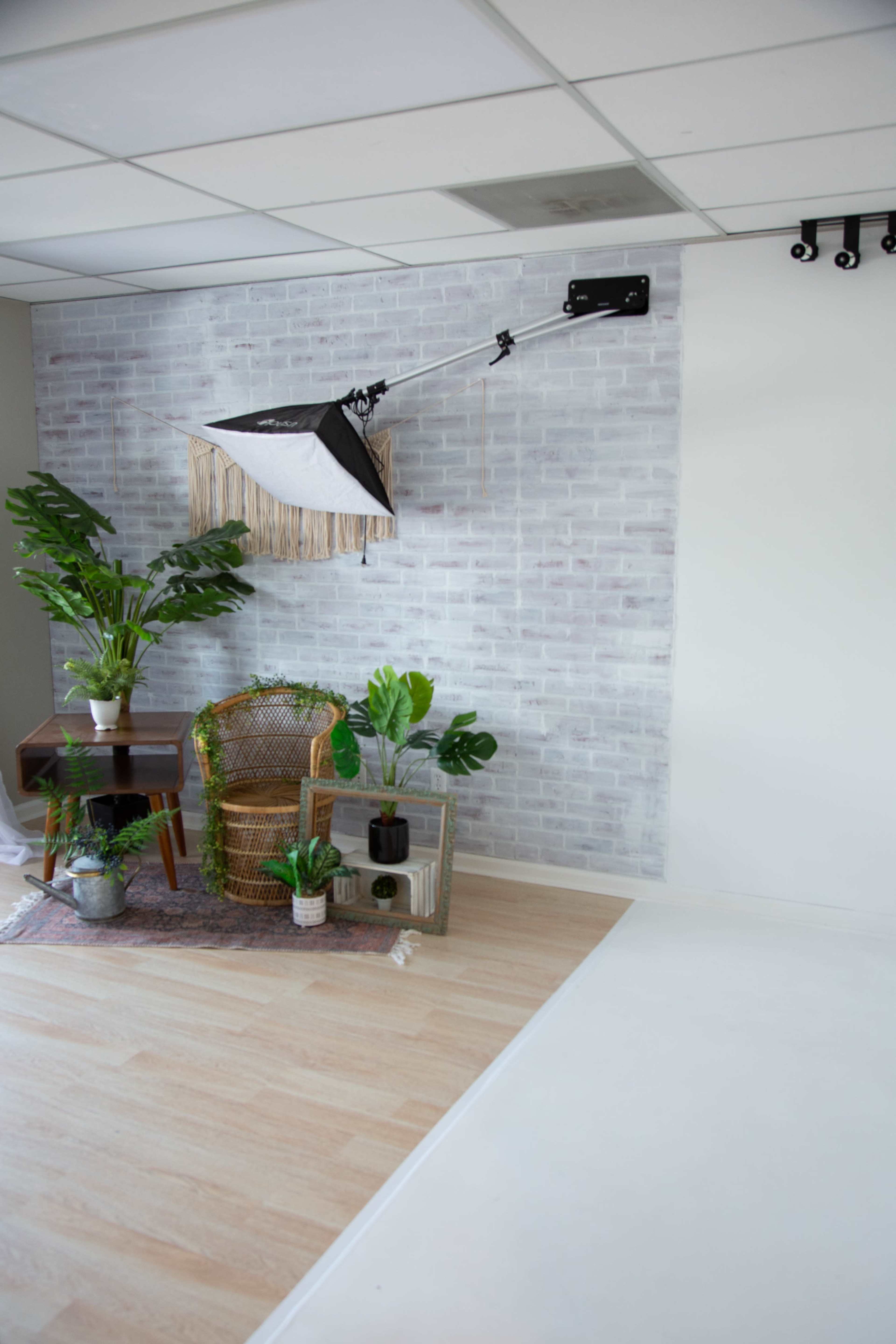 A minimalistic interior space featuring a woven chair, a table with plants, and a light reflector mounted on the ceiling against a brick-patterned wall.