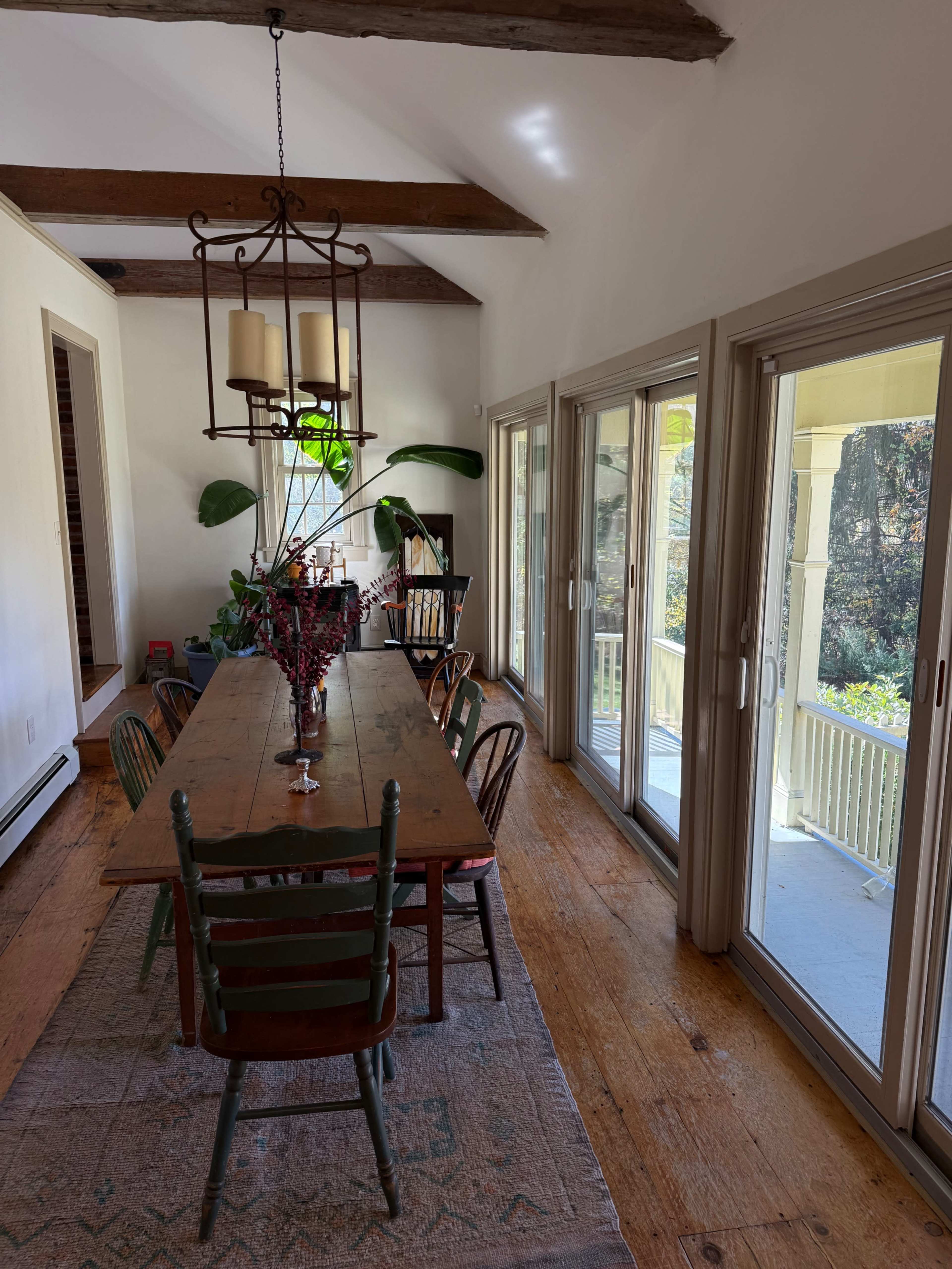 The image shows a dining area with a long wooden table surrounded by chairs, large windows, and a view of an outdoor porch.