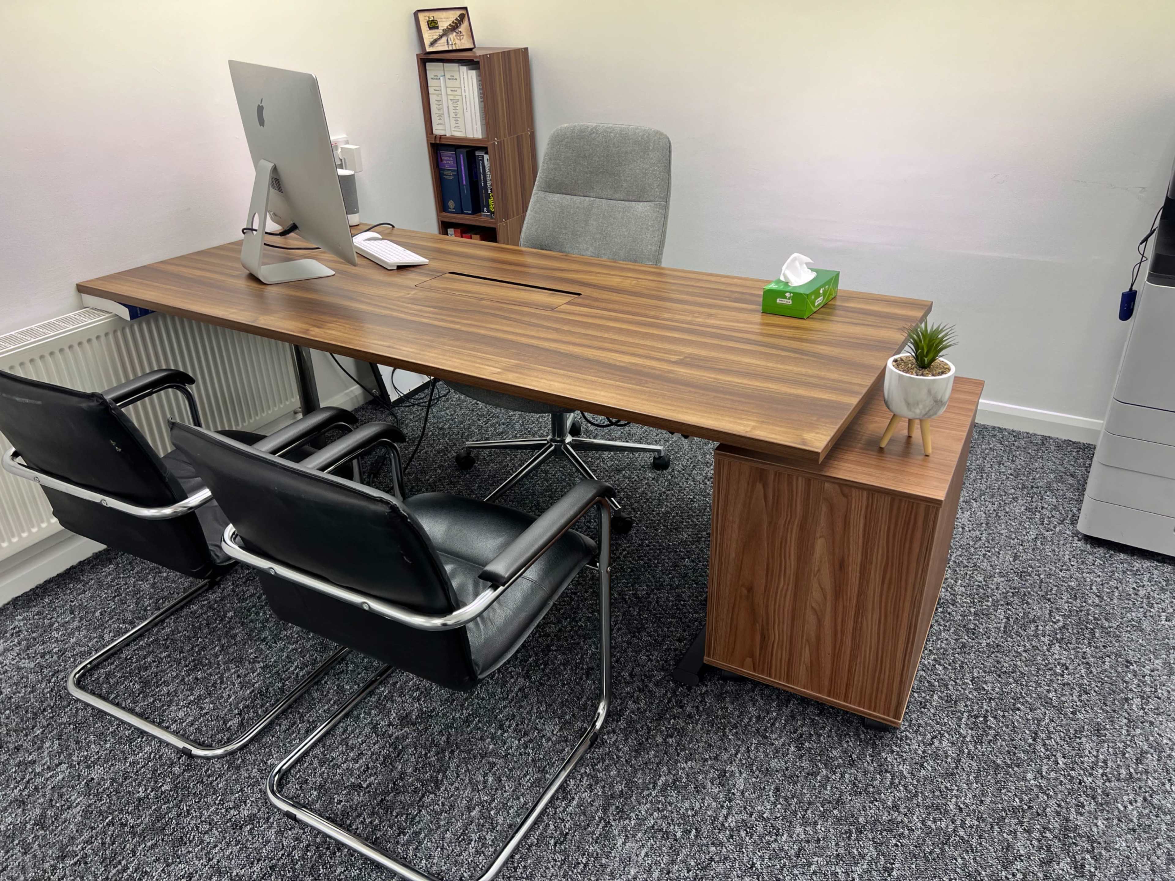The image shows a modern office space with a wooden desk, an all-in-one iMac computer, two black chairs, and a small plant on the desk.