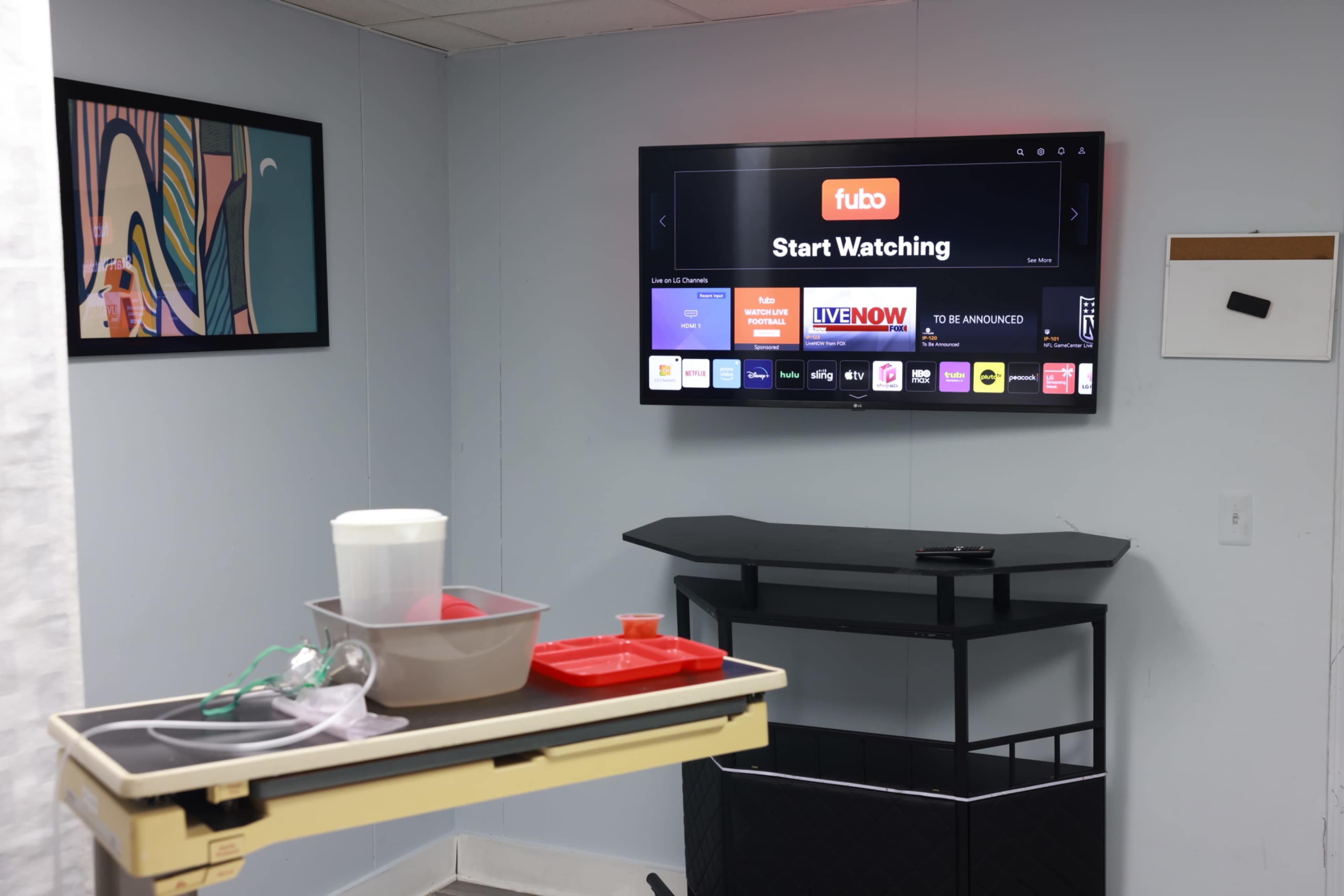 The image shows a small room with a flat-screen TV mounted on the wall displaying a streaming service interface, alongside a cart with medical supplies and a framed artwork.