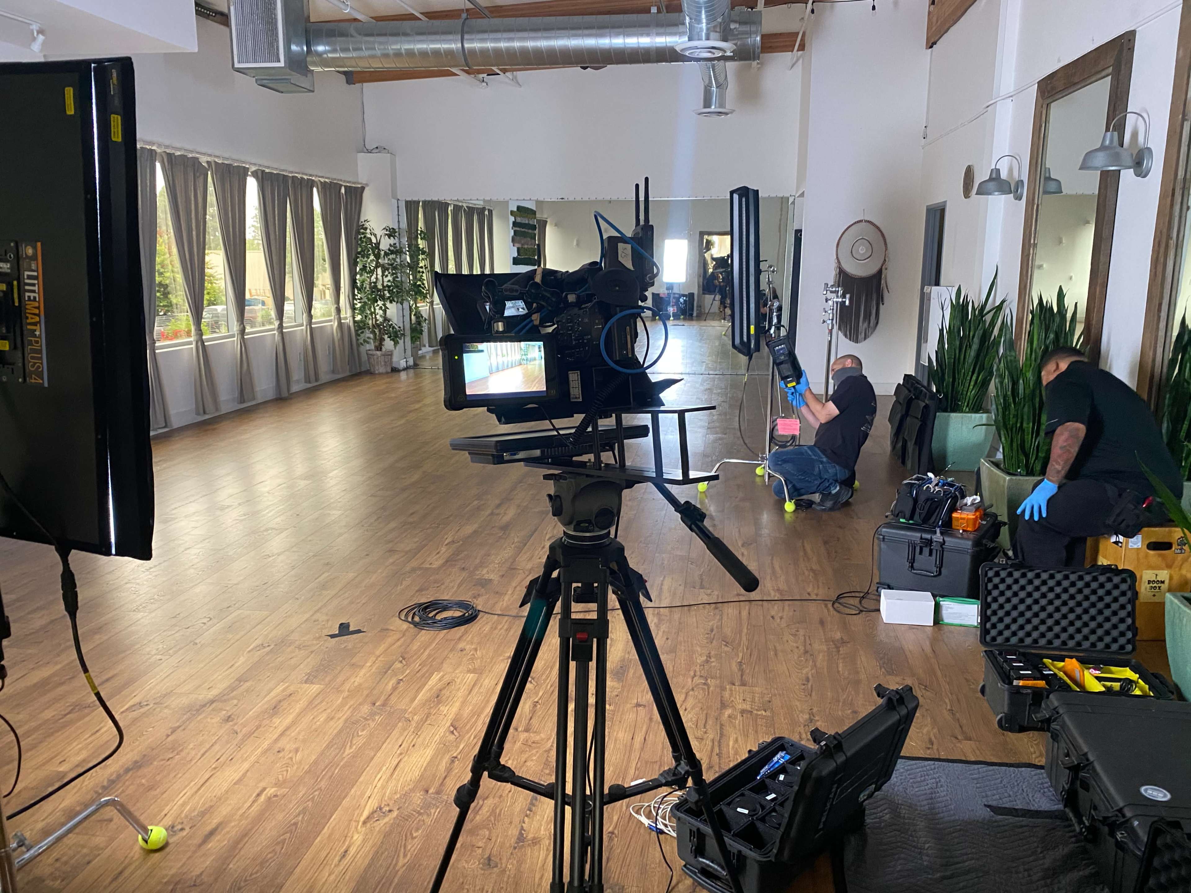 A camera setup is positioned in a spacious studio with wooden floors, while crew members prepare equipment in the background.