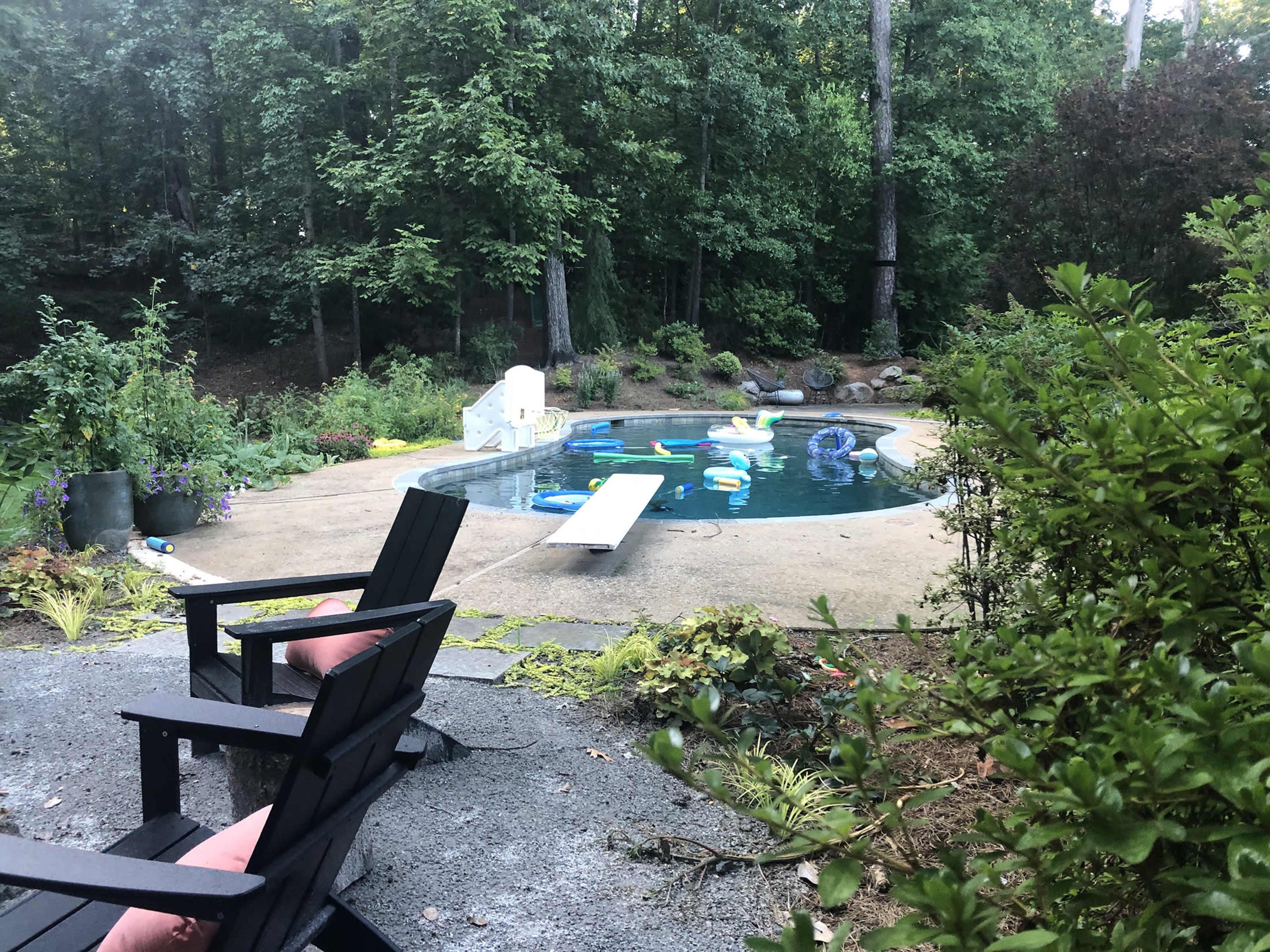 A pool with colorful floatation devices is surrounded by greenery and chairs on a stone pathway.