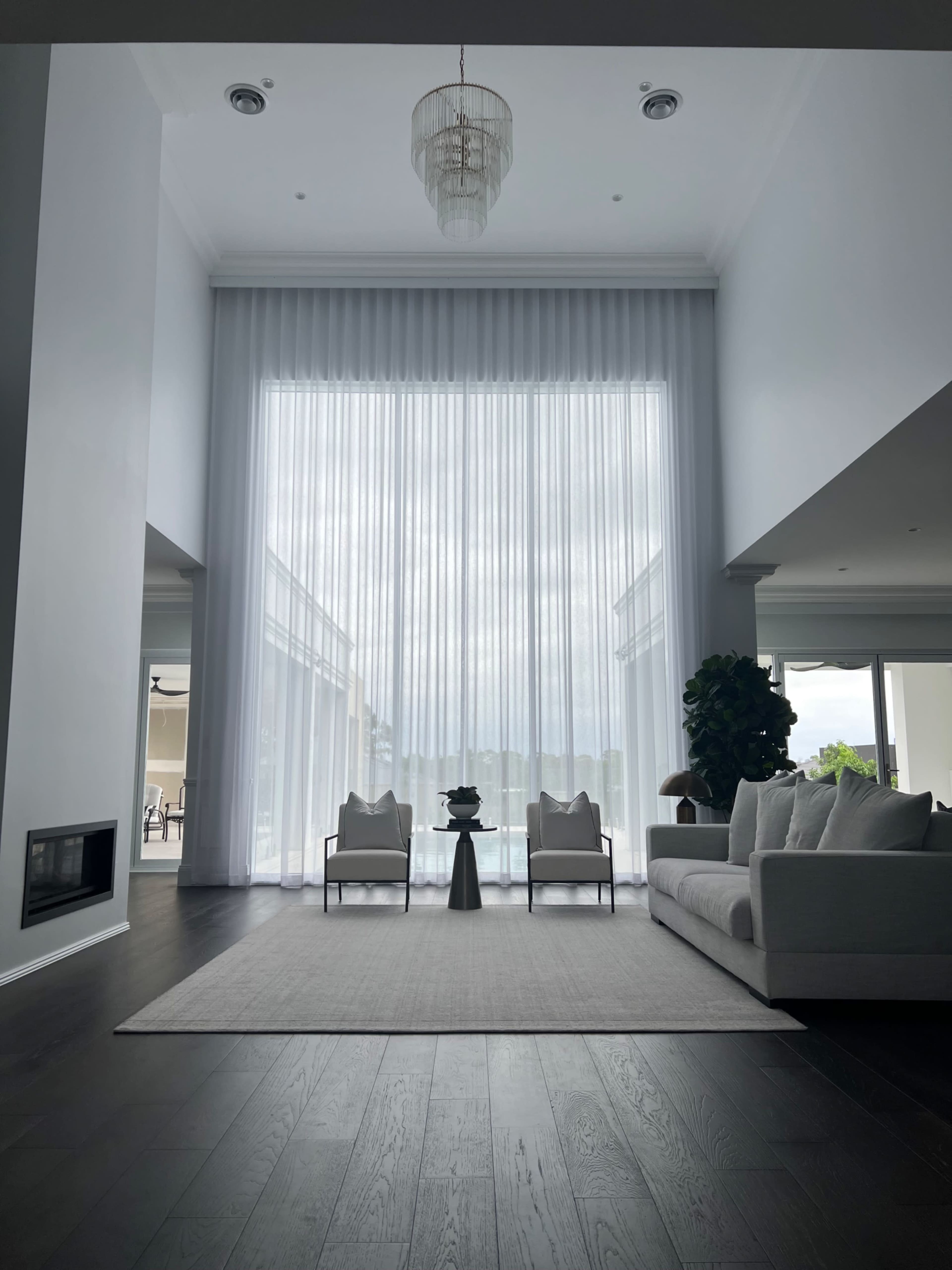 The image shows a spacious living room with large sheer curtains, two armchairs, a decorative table, and a view of an outdoor pool area through large windows.