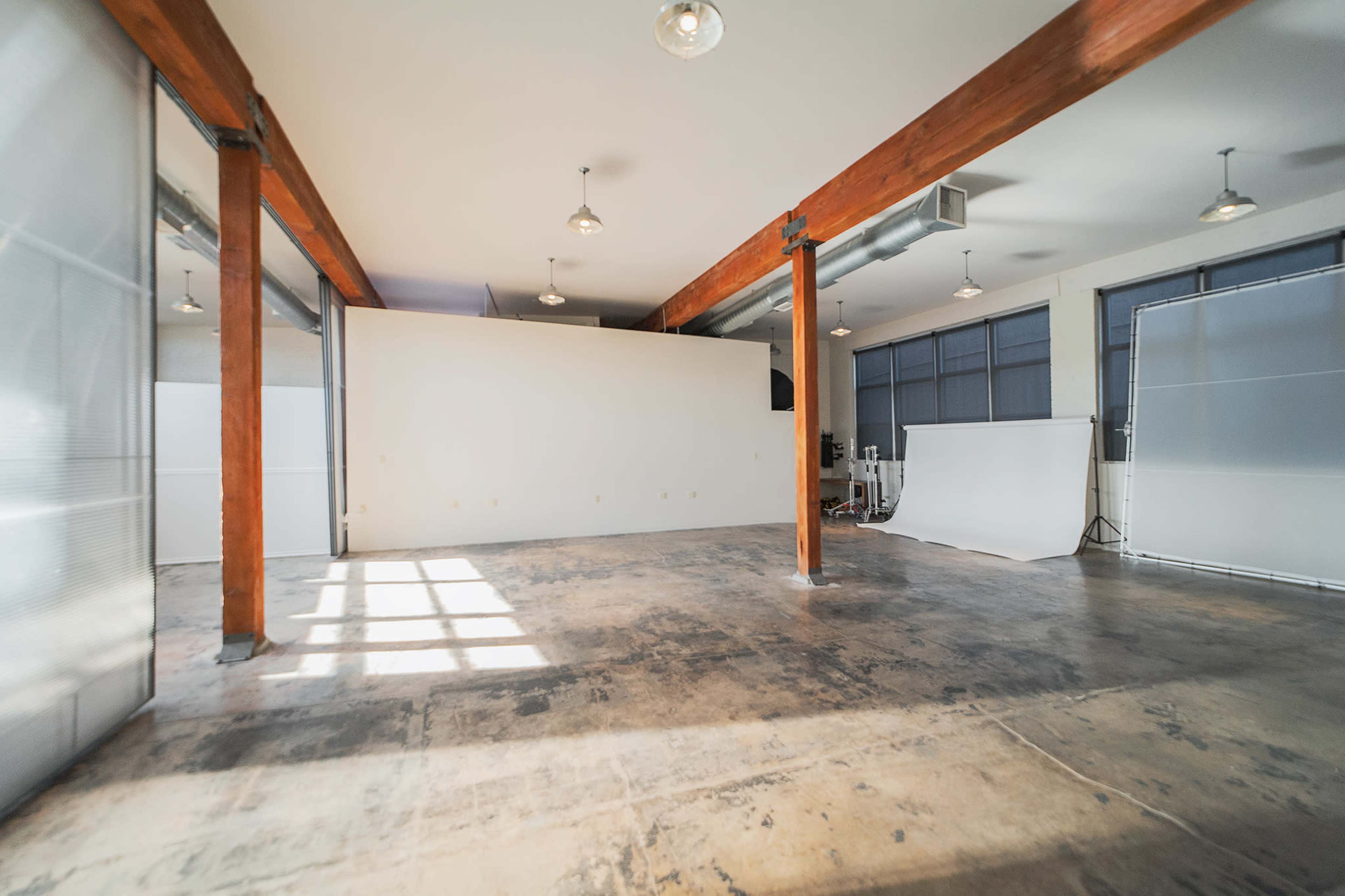 A spacious, empty room with wooden beams and large windows, featuring a light-colored wall and a white backdrop at one end.