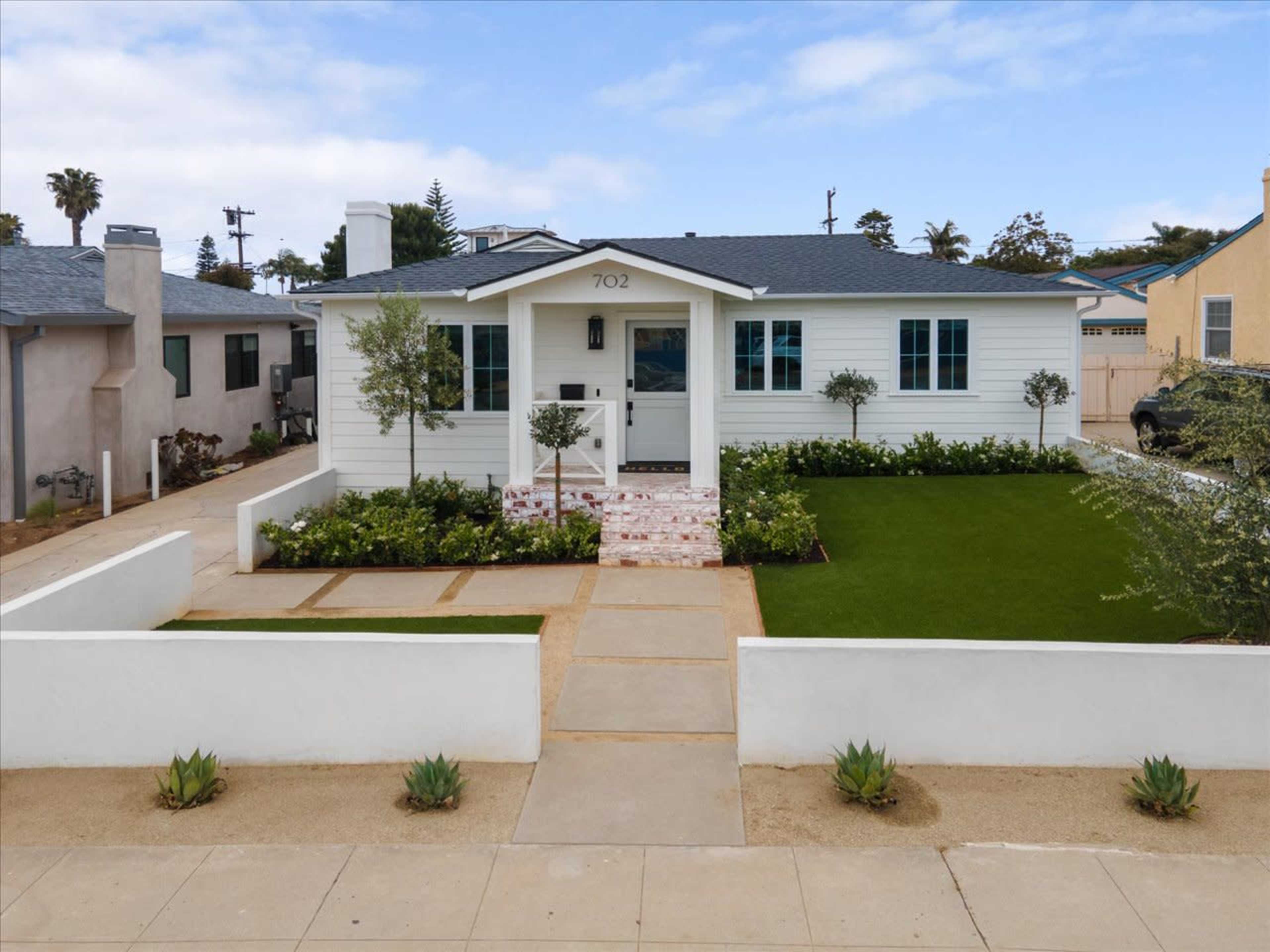 A single-story white house with a front lawn, bordered by a low wall and a walkway leading to the main entrance.