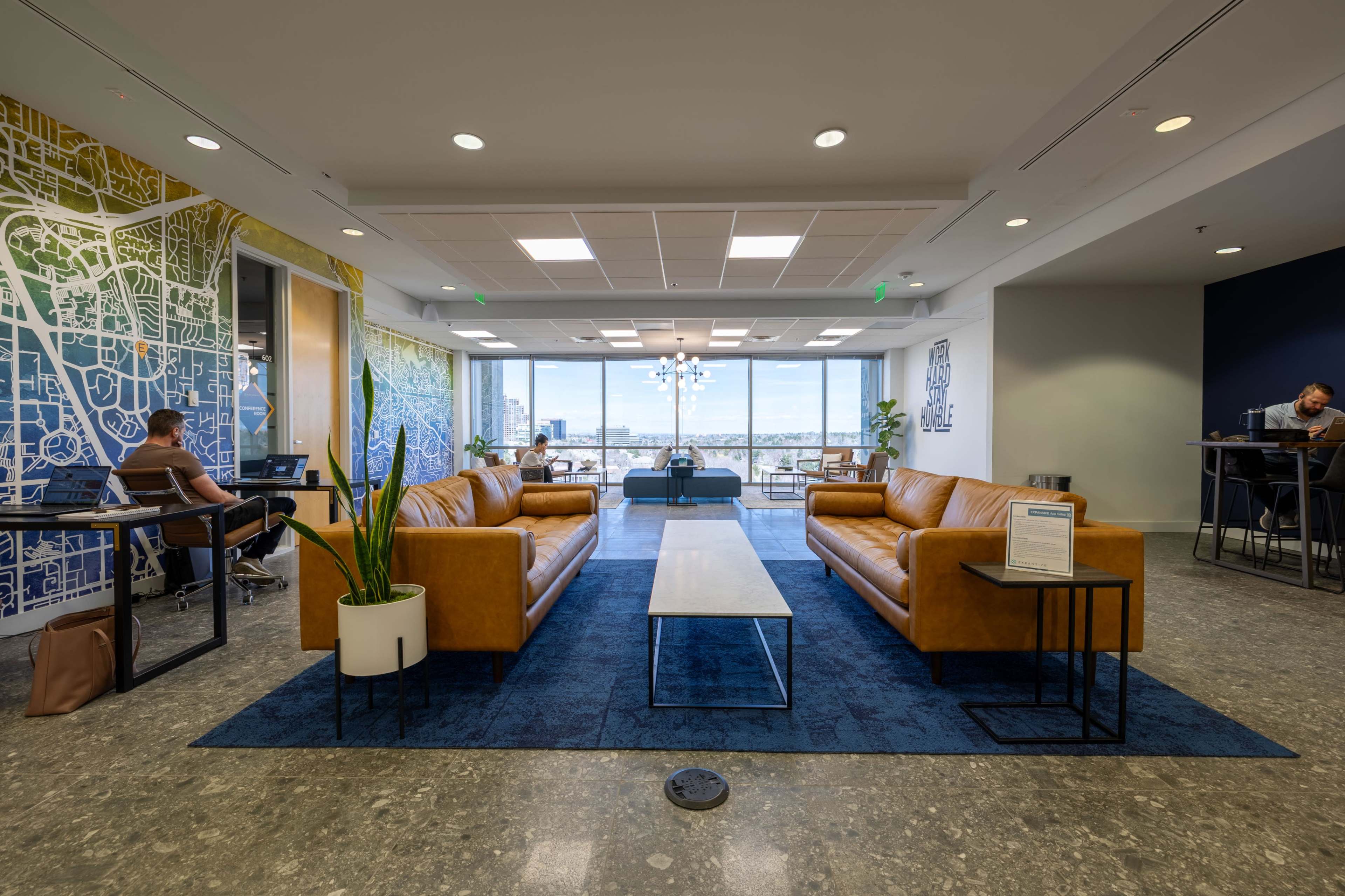 The image shows a modern office lounge area featuring brown leather sofas, a coffee table, and large windows with a view, along with individuals working at desks.