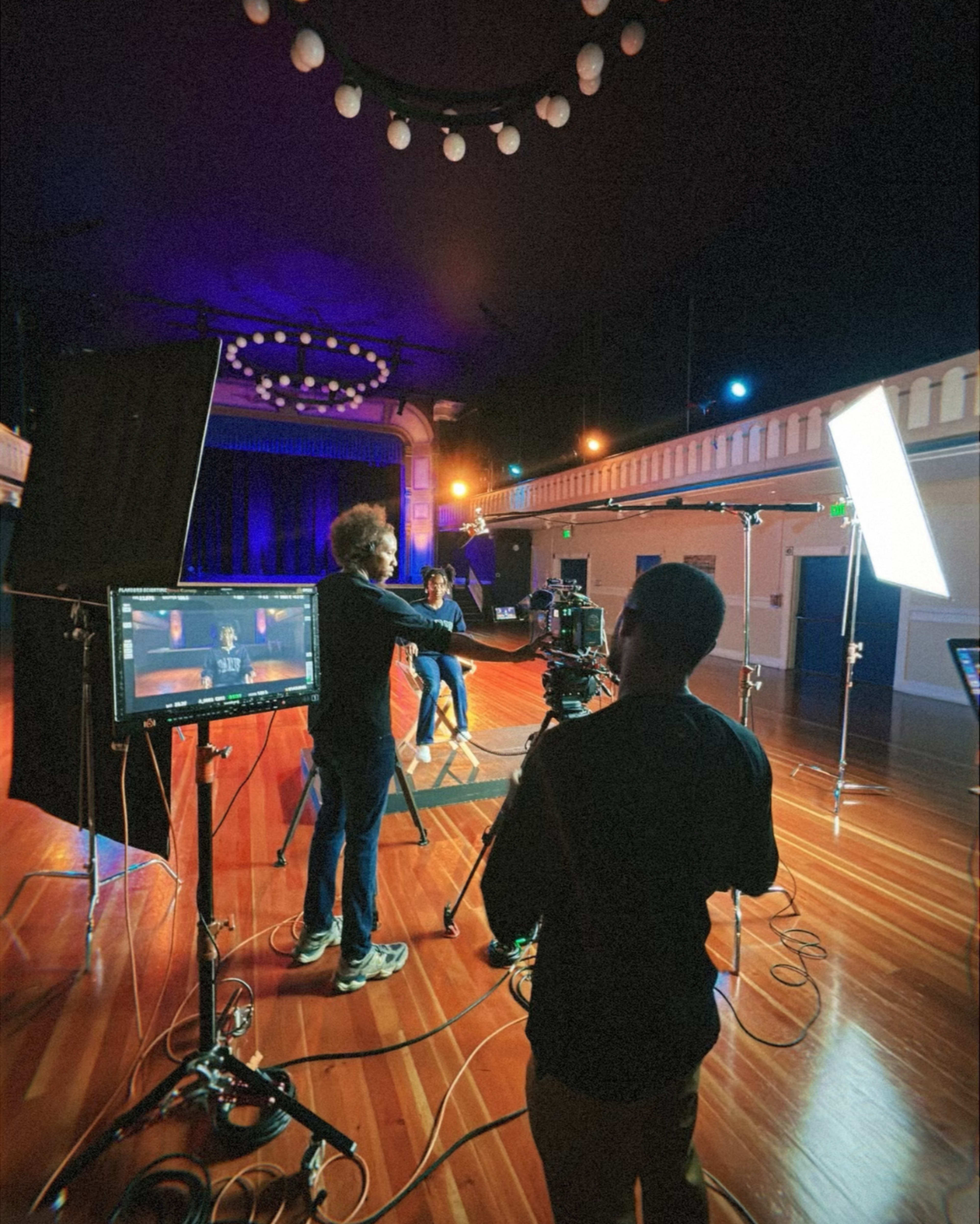 A camera crew sets up equipment in a spacious, dimly lit auditorium for a video shoot.