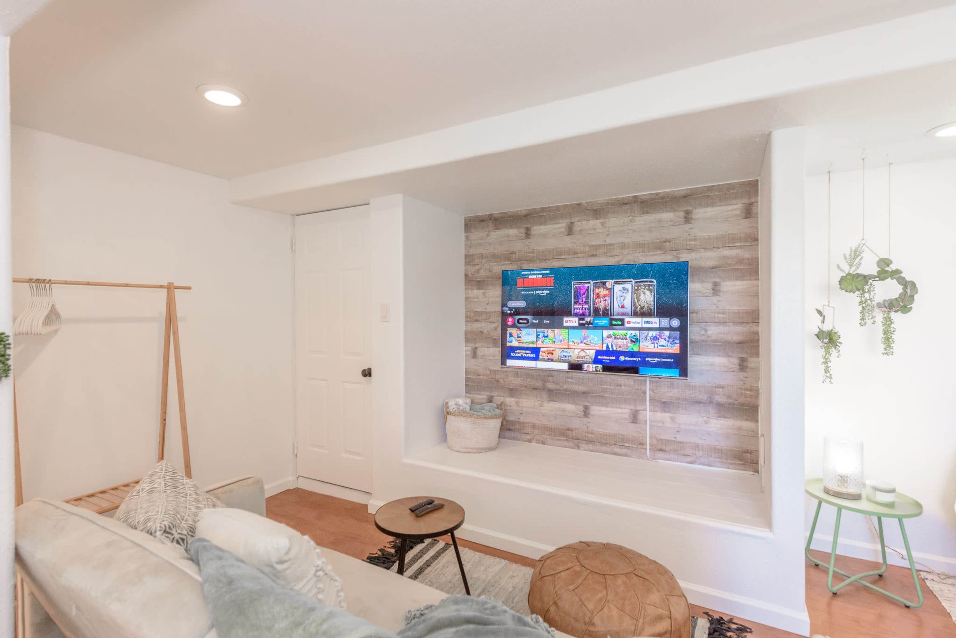 A modern living space featuring a light-colored sofa, a wall-mounted television set against a wooden backdrop, and minimalist decor including a small side table and plants.