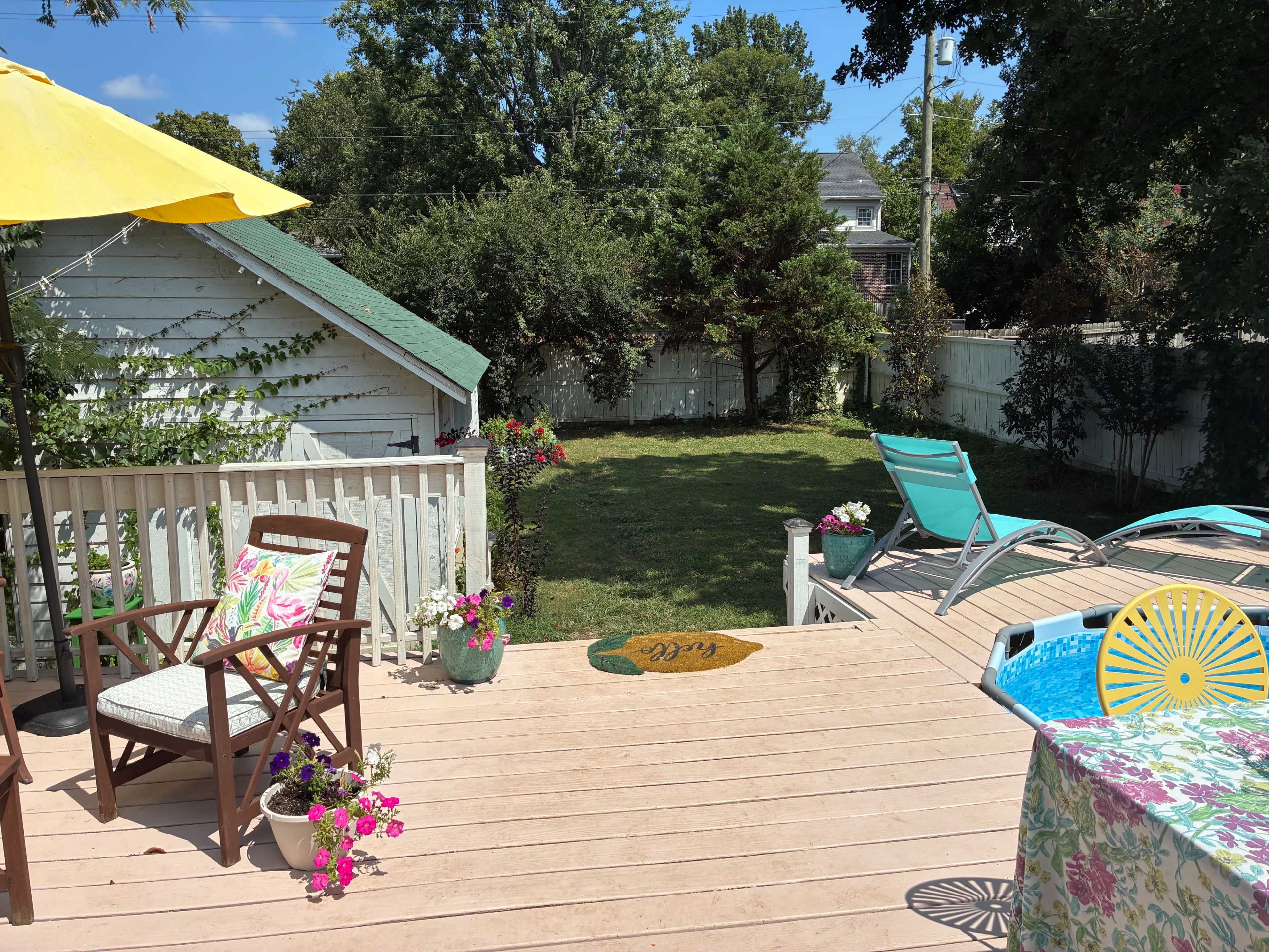 The image shows a backyard deck area with a seating arrangement, an umbrella, and lawn chairs overlooking a green lawn and surrounding plants.