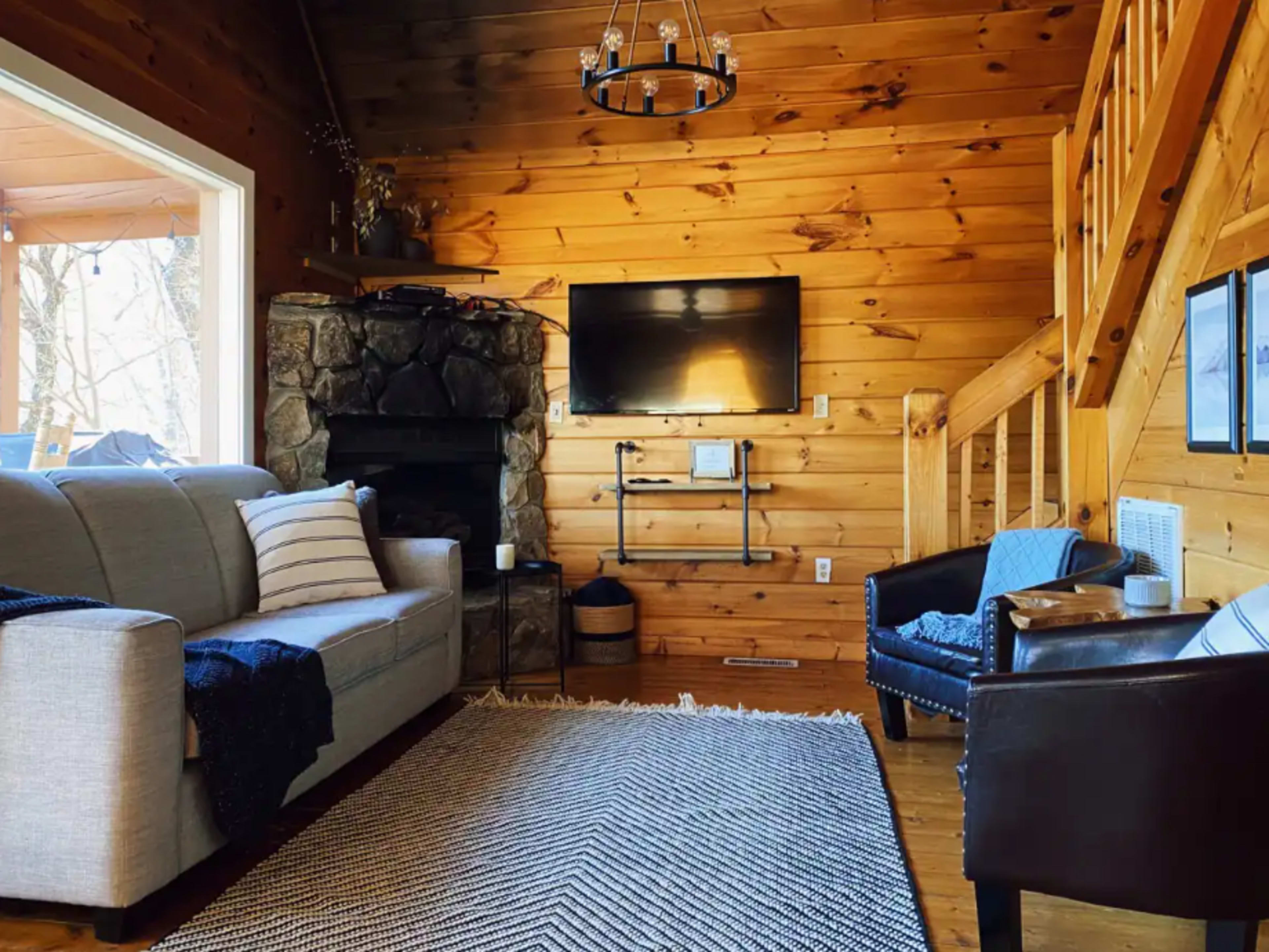 The living room features a gray sofa, a stone fireplace, a mounted television, wooden walls, and a staircase.