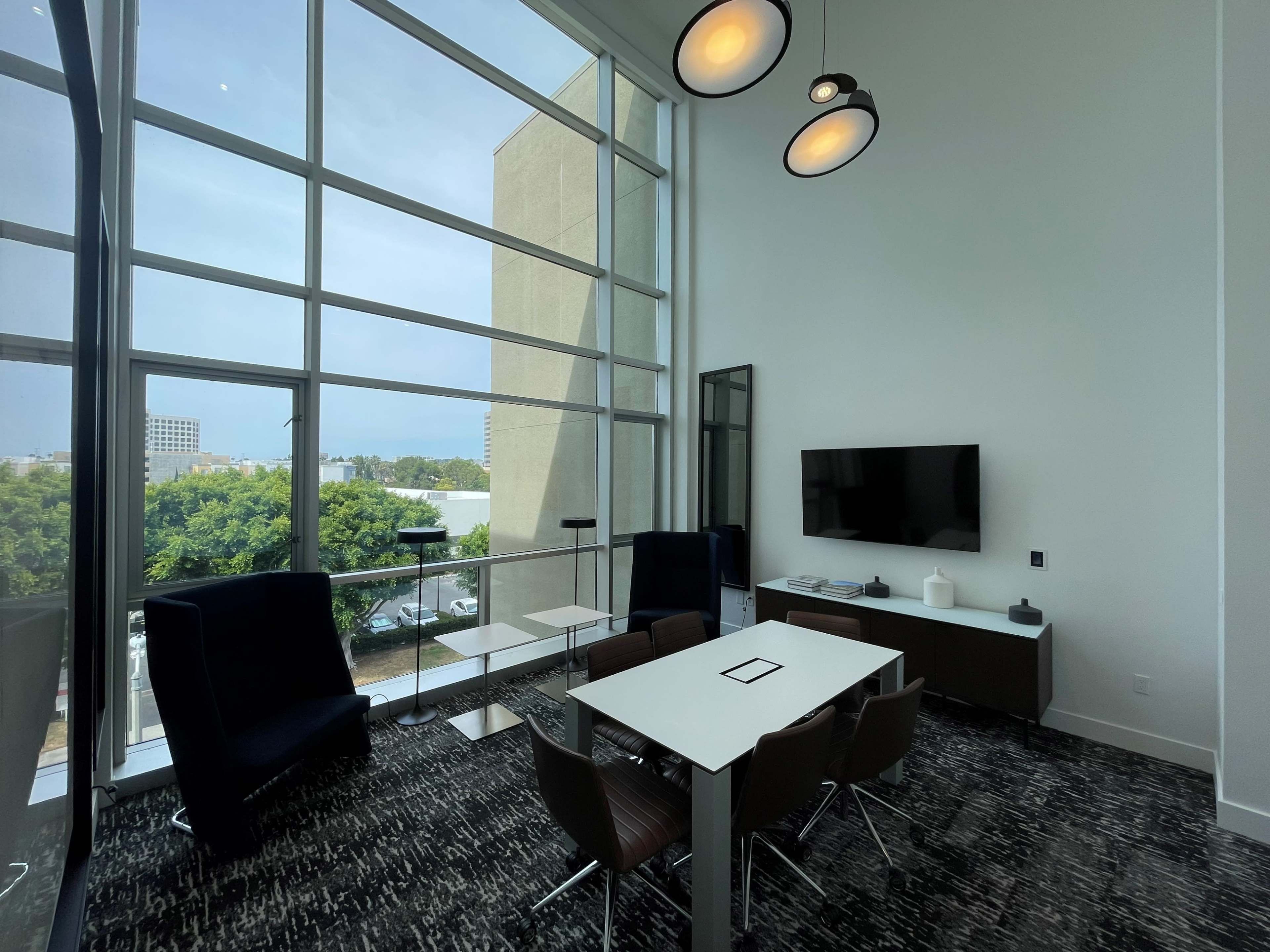 A modern meeting room with large windows, a rectangular table, chairs, and a television mounted on the wall, overlooking a green area outside.