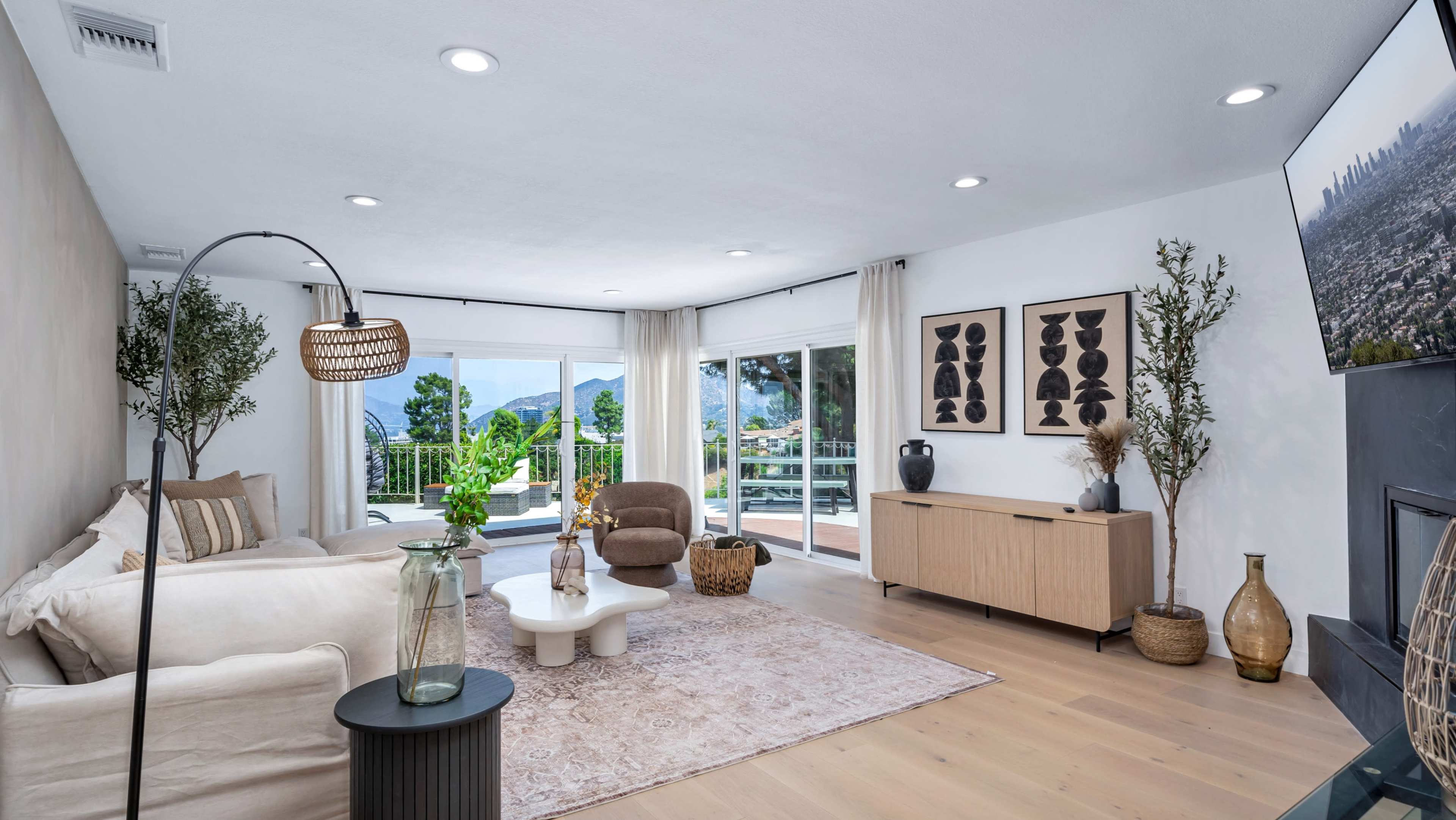The image shows a modern living room featuring a light-colored sofa, a round coffee table, a wooden sideboard, and large windows overlooking a green landscape.