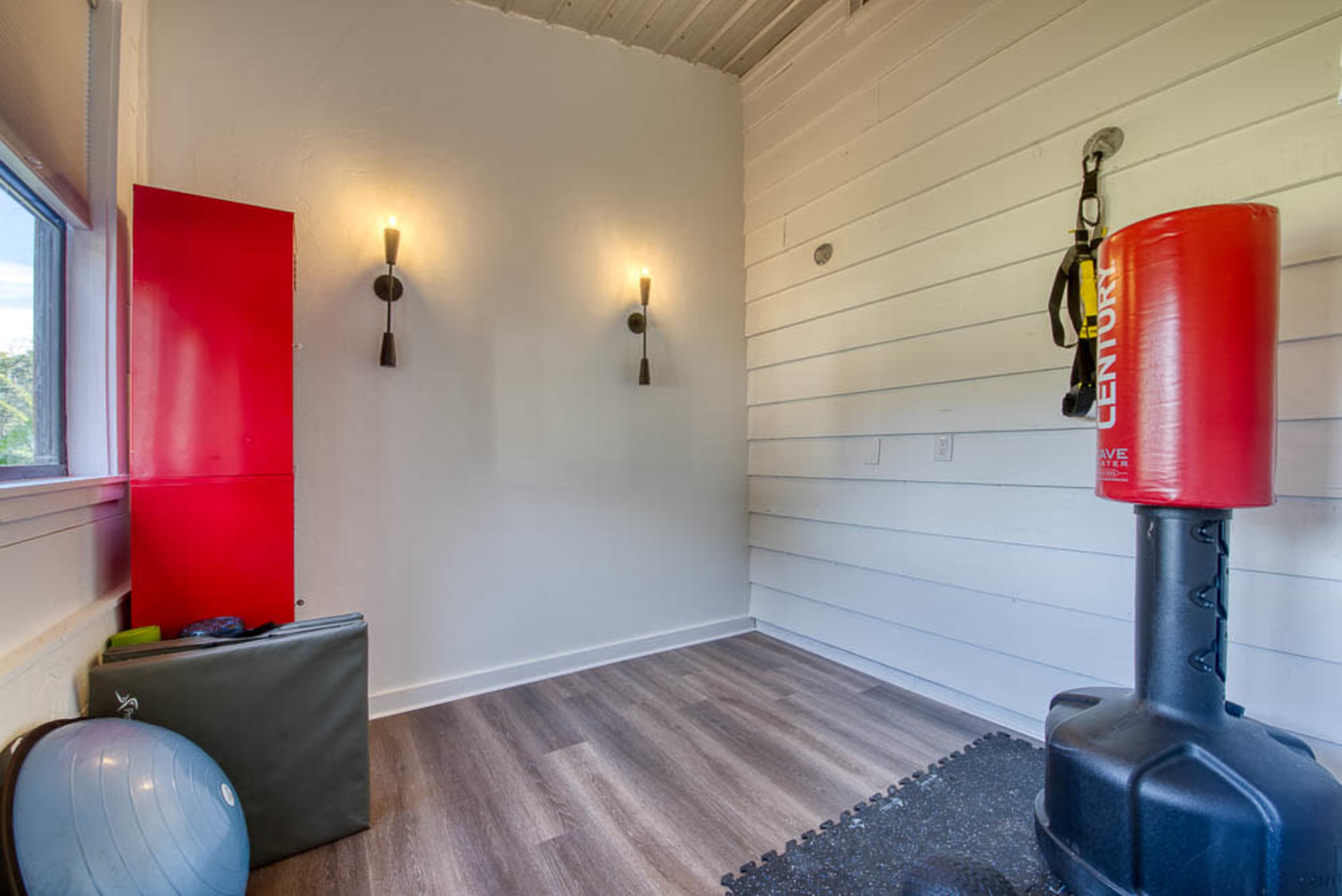 A minimalistic home gym with a punching bag, exercise ball, and wall-mounted lights against a white shiplap wall.