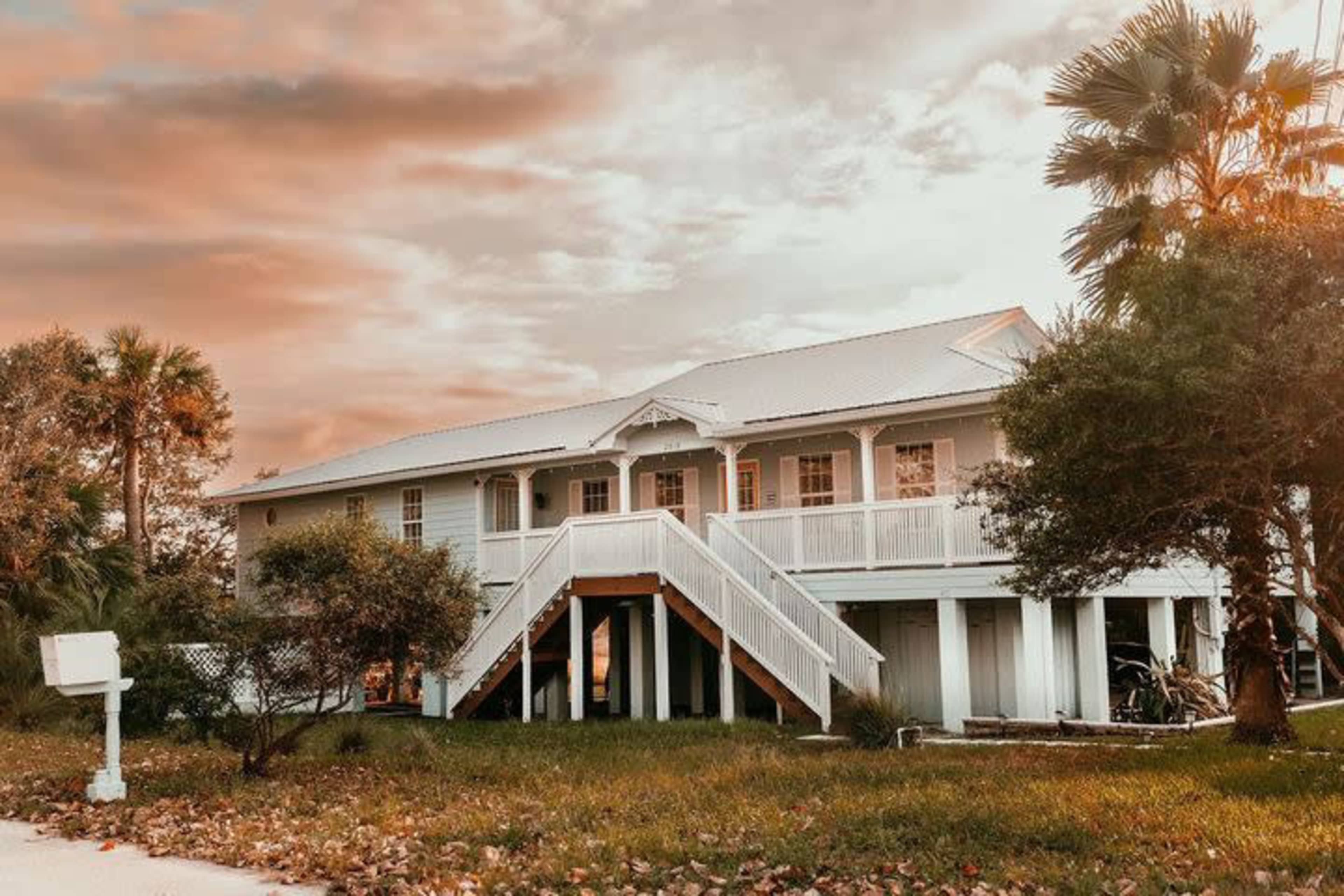 A two-story white wooden house with a staircase leading to the entrance is surrounded by palm trees and grassy landscaping.