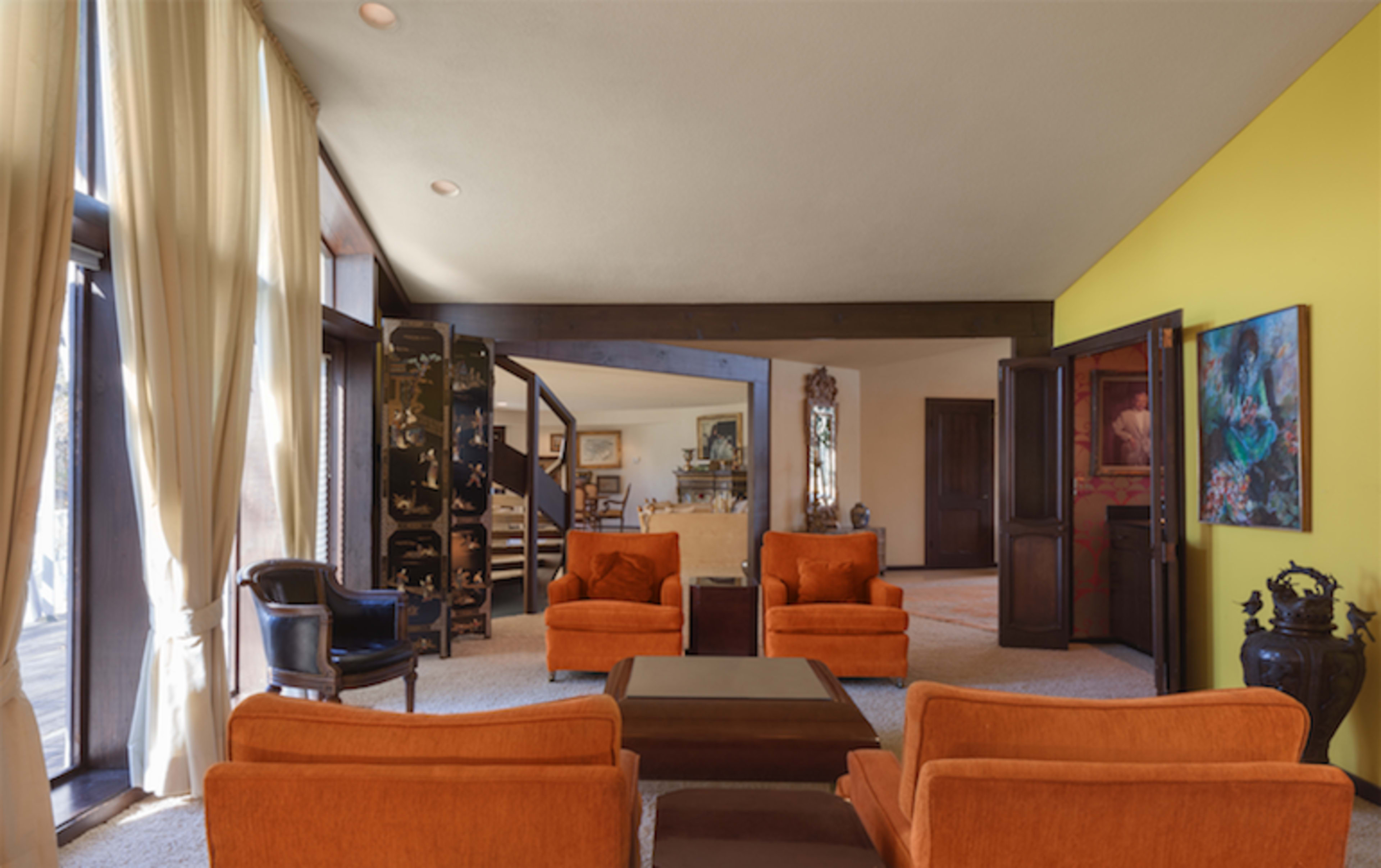 The image shows a spacious living room featuring orange armchairs, a coffee table, and large windows that allow natural light to enter, with a bright yellow accent wall.
