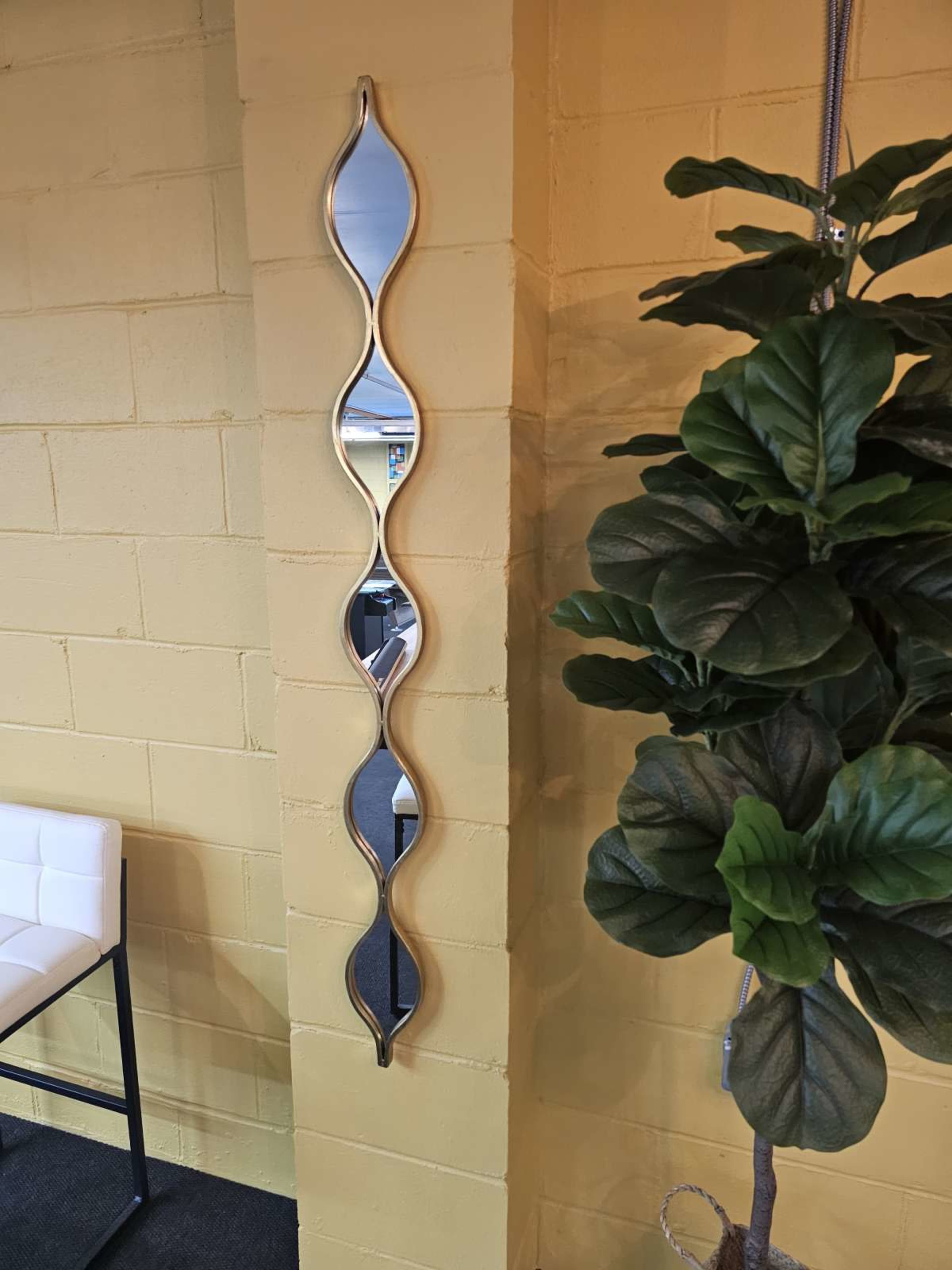 The image shows a narrow, wavy mirror mounted on a yellow wall beside a large green indoor plant.