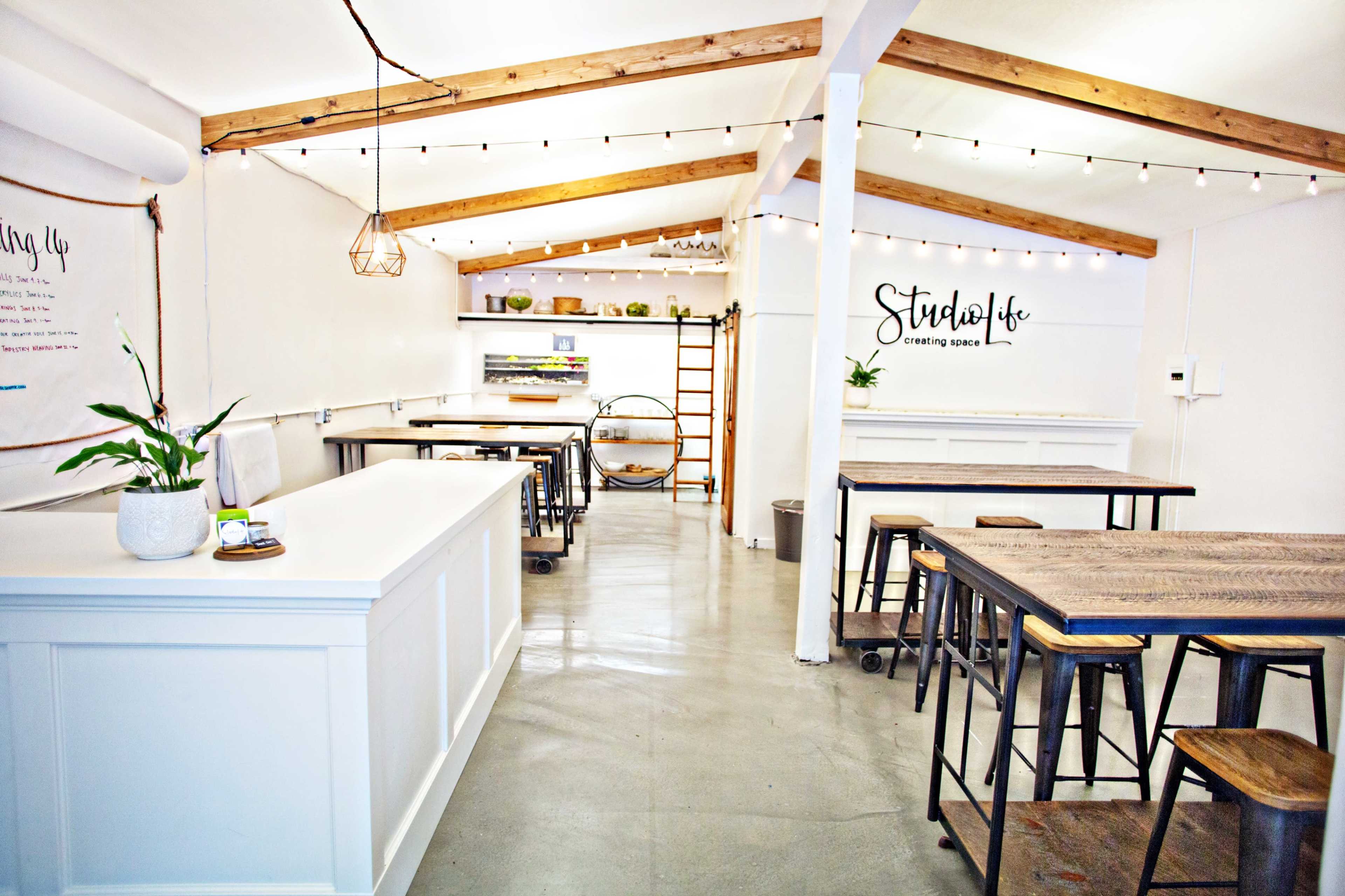 A bright, minimalist workspace with white walls, wooden beams, and a series of tables and stools arranged along the room.