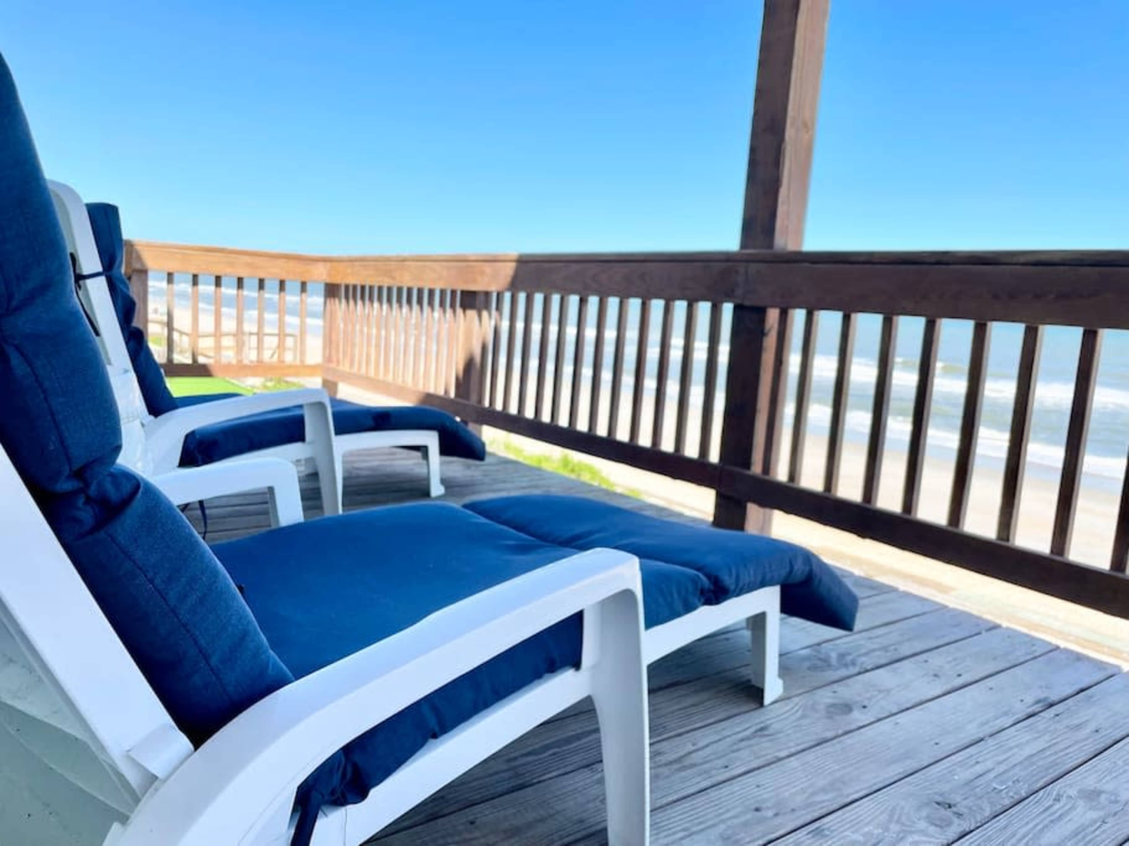 Lounge chairs with blue cushions are arranged on a wooden deck overlooking the ocean waves.