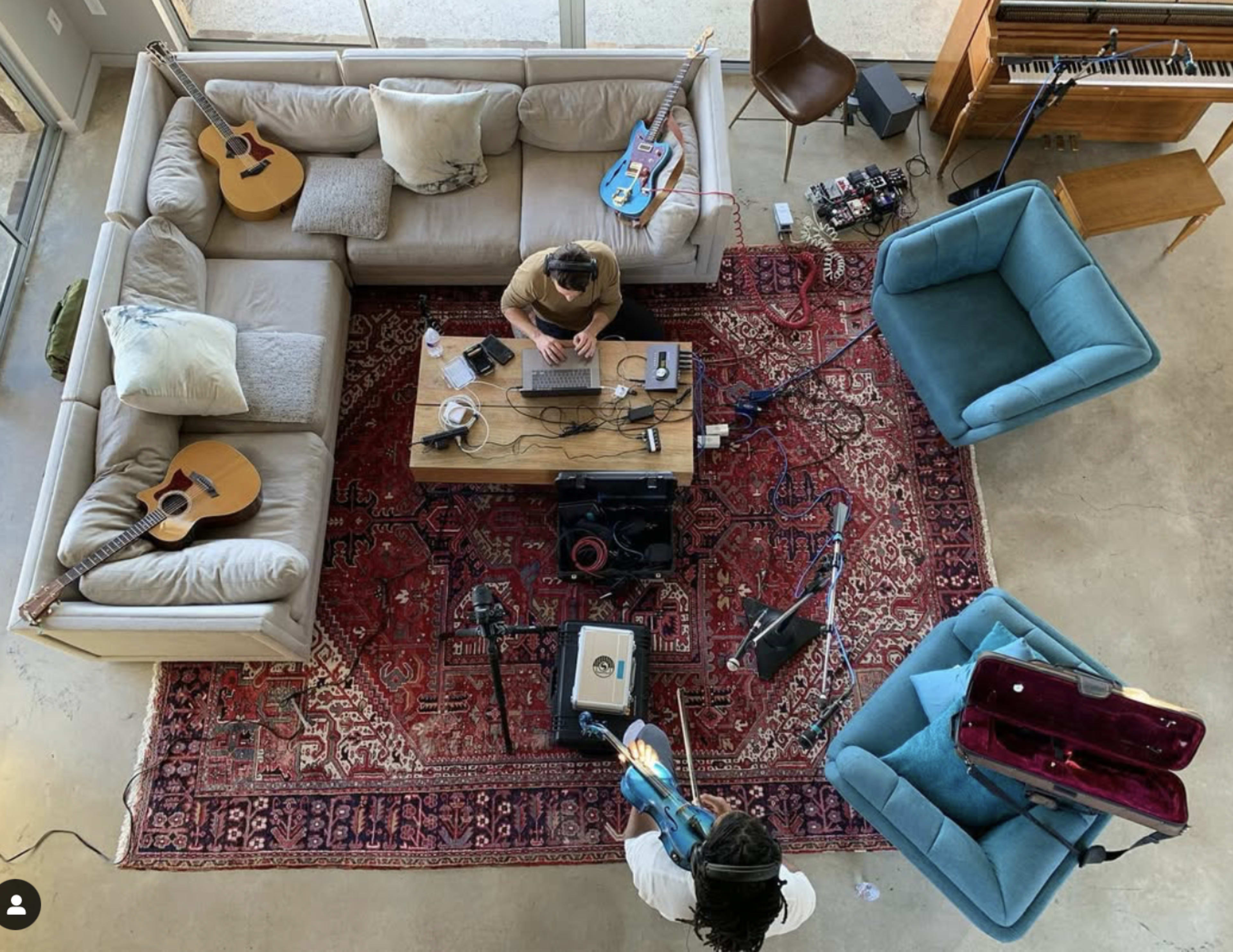 A person sits at a table with a laptop and musical equipment in a cozy living room featuring multiple guitars and an upright piano.