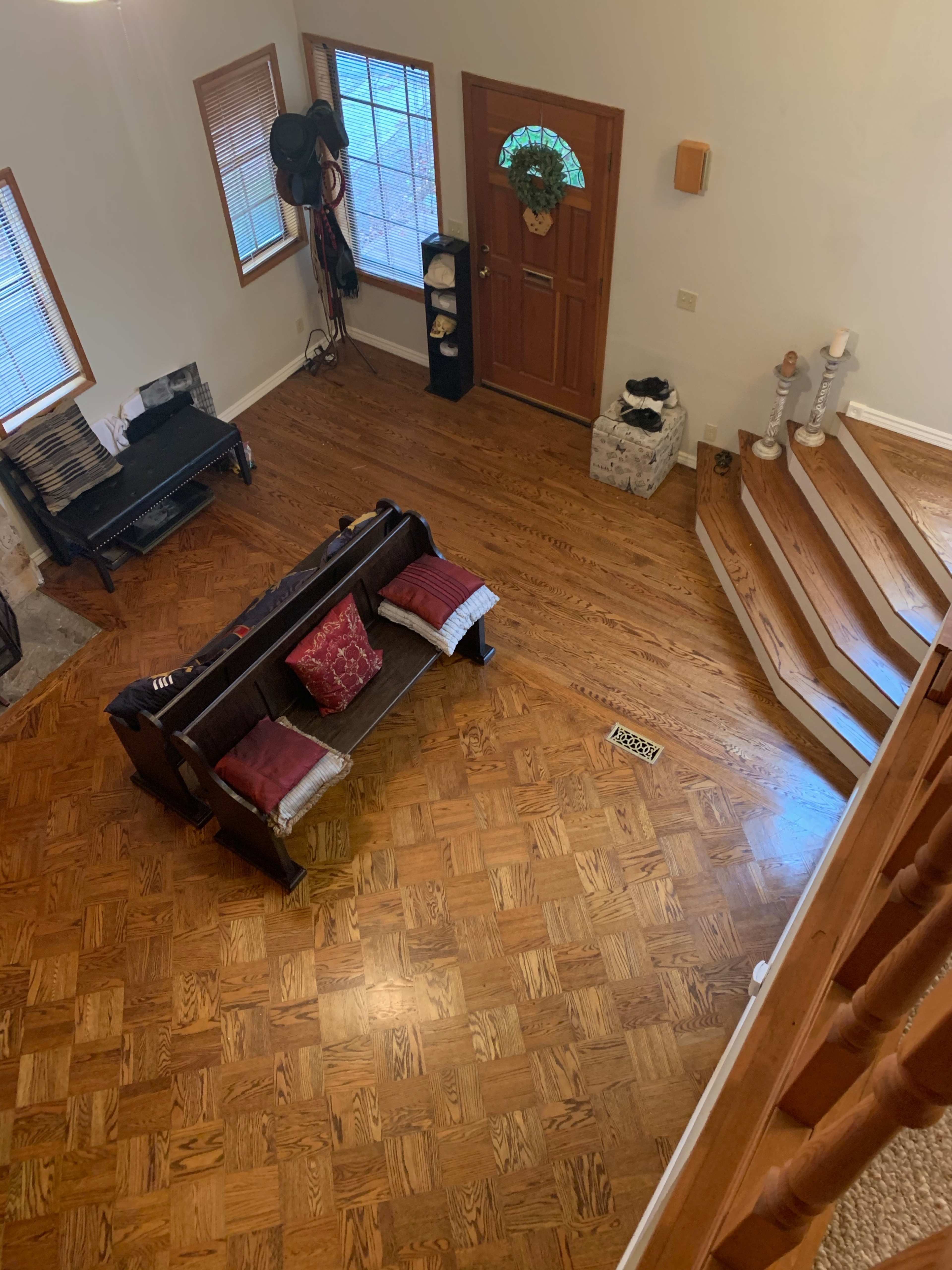The image shows a living room with a wooden floor, a dark couch with red cushions, a staircase, and a front door with a wreath.