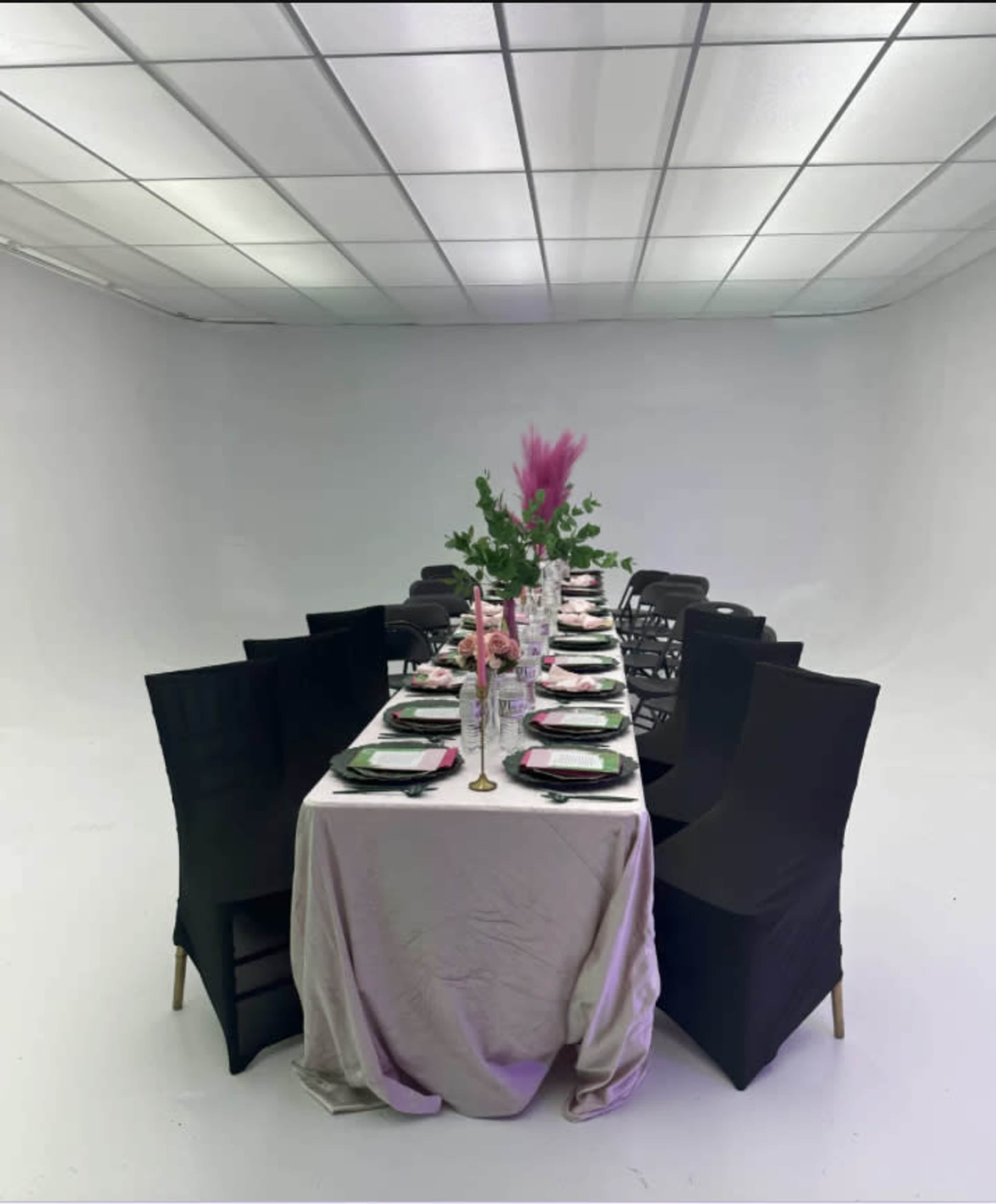 A long dining table is set with black plates, glasses, and floral arrangements, surrounded by black chairs in a stark white room.