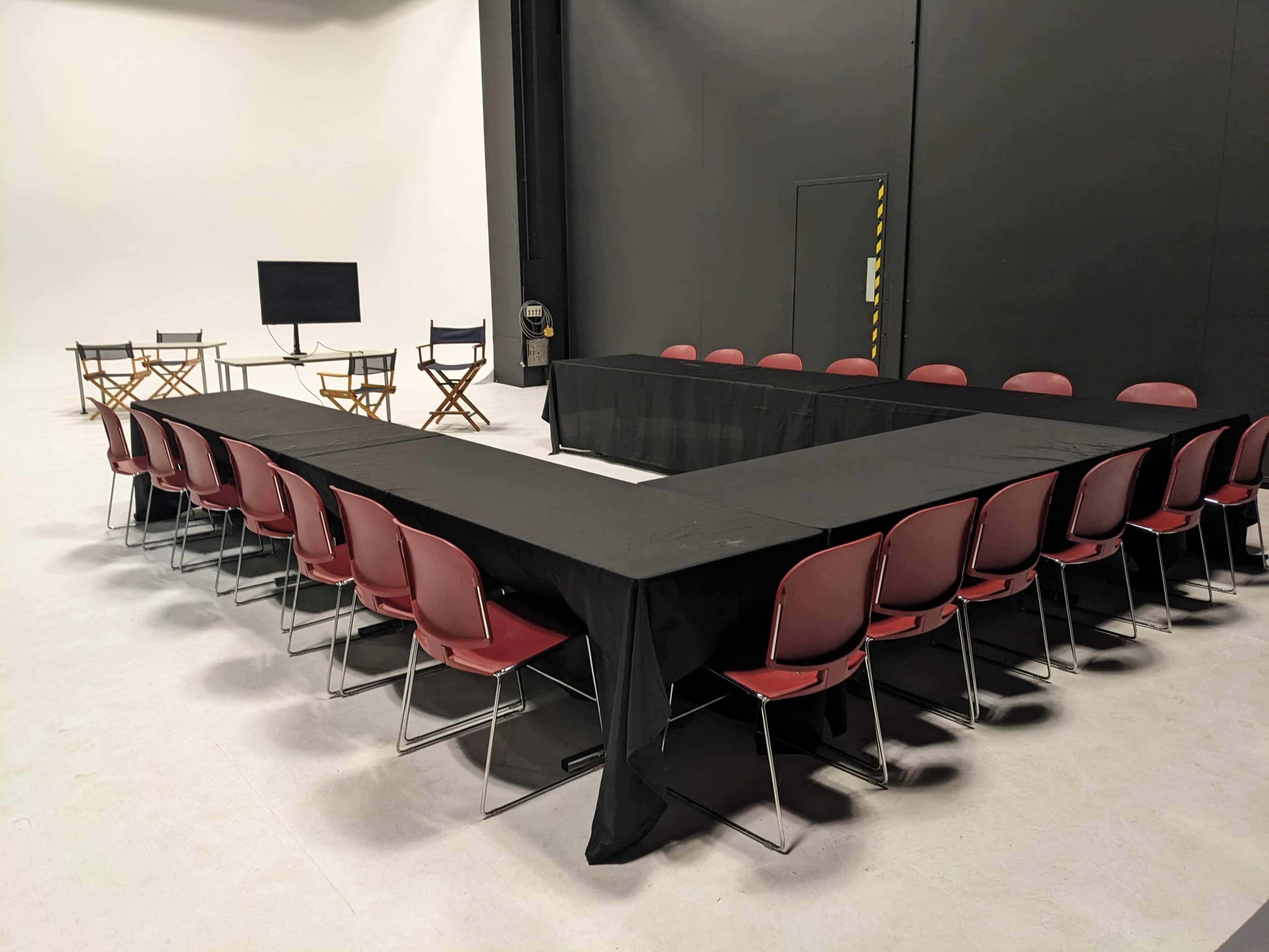 A rectangular table arrangement with red chairs is set up in a studio space, with two additional chairs and a television screen in the background.