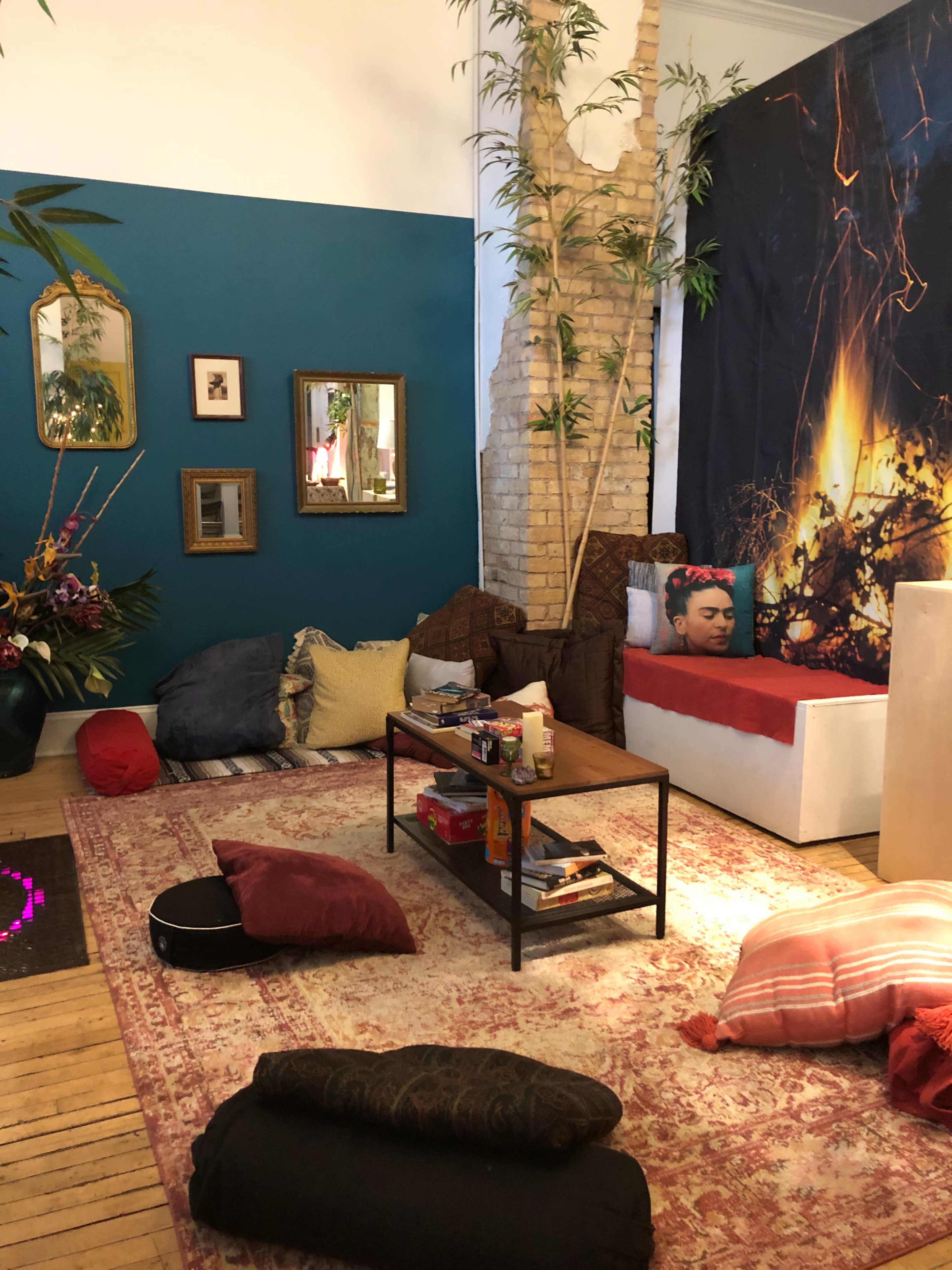 The room features a mix of seating options including pillows and a low table, with a red bedspread and a wall adorned with plants and artwork.