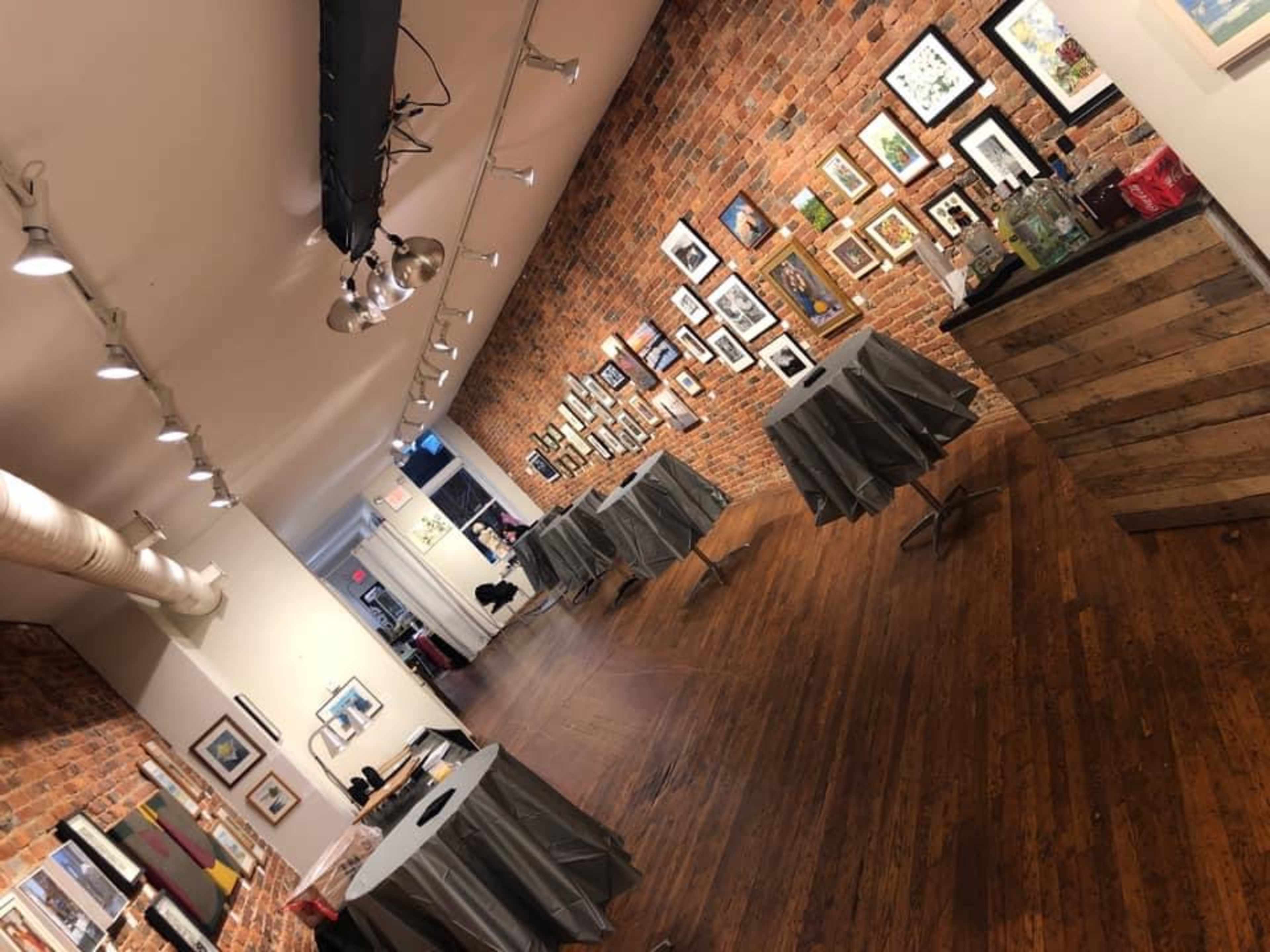 An art gallery with brick walls, wooden flooring, and tables covered with gray cloths, displaying framed artworks on the walls.