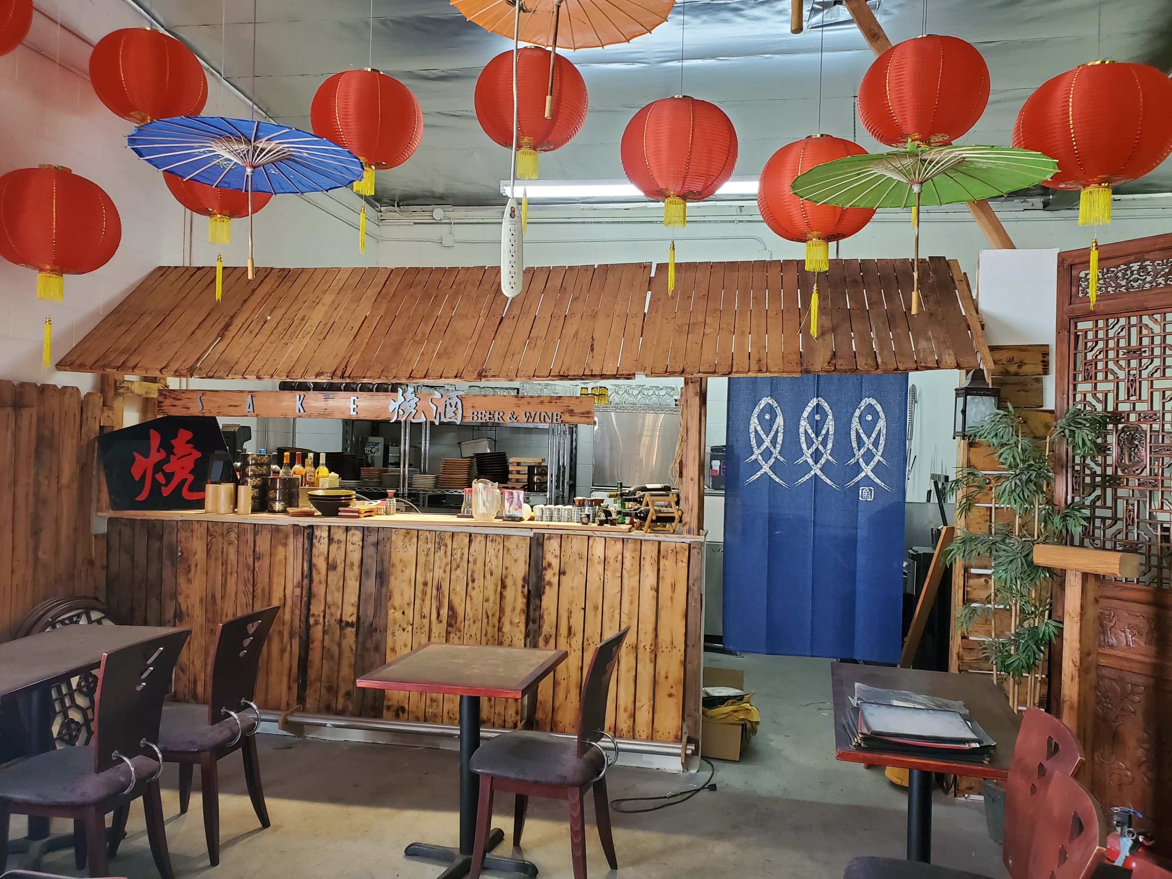A wooden counter with hanging red lanterns and colorful paper umbrellas is seen in an Asian-themed restaurant.