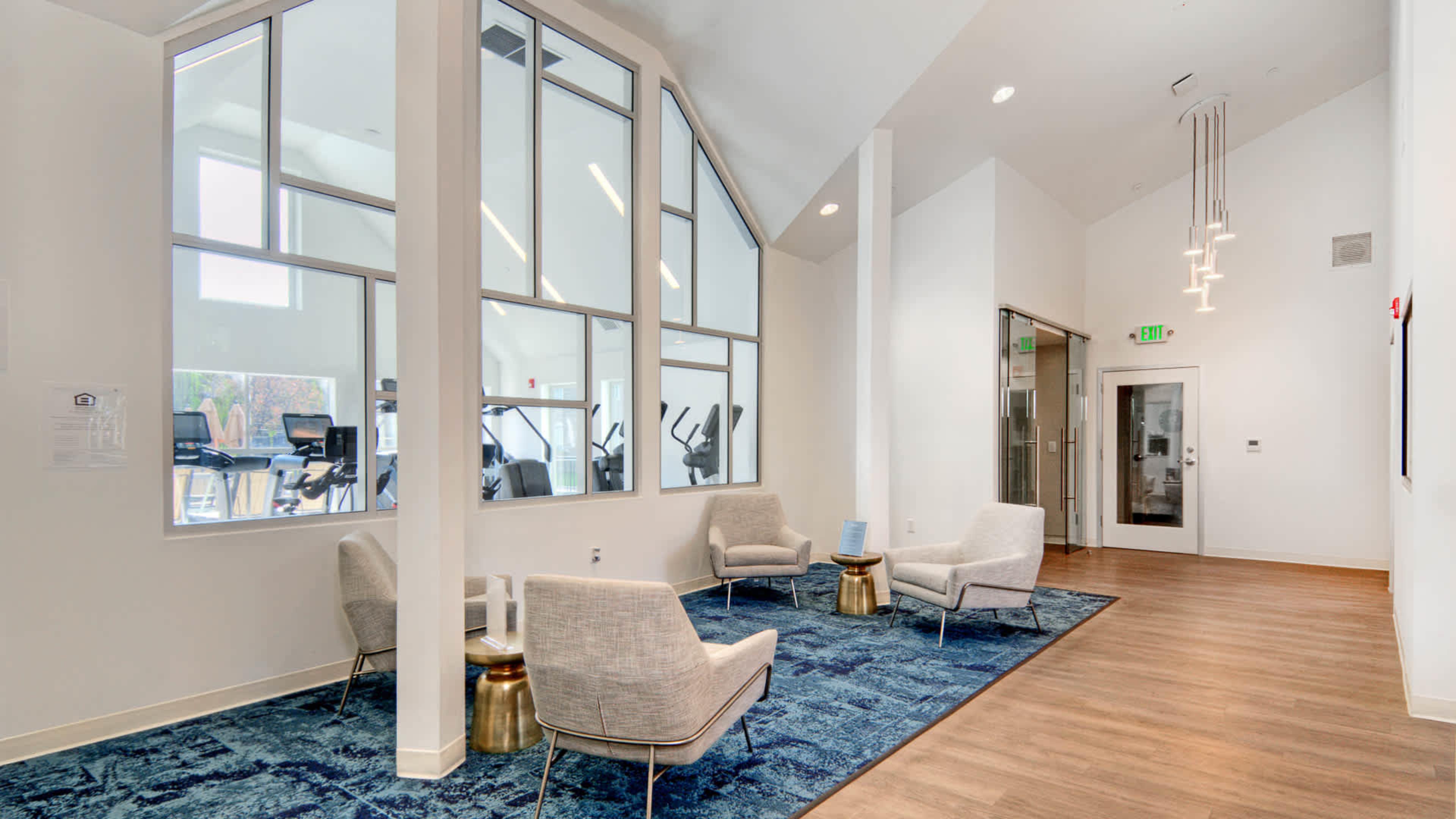 A modern lobby area with light-colored walls, two seating arrangements of gray chairs on a blue carpet, and large windows providing an view of a fitness area.