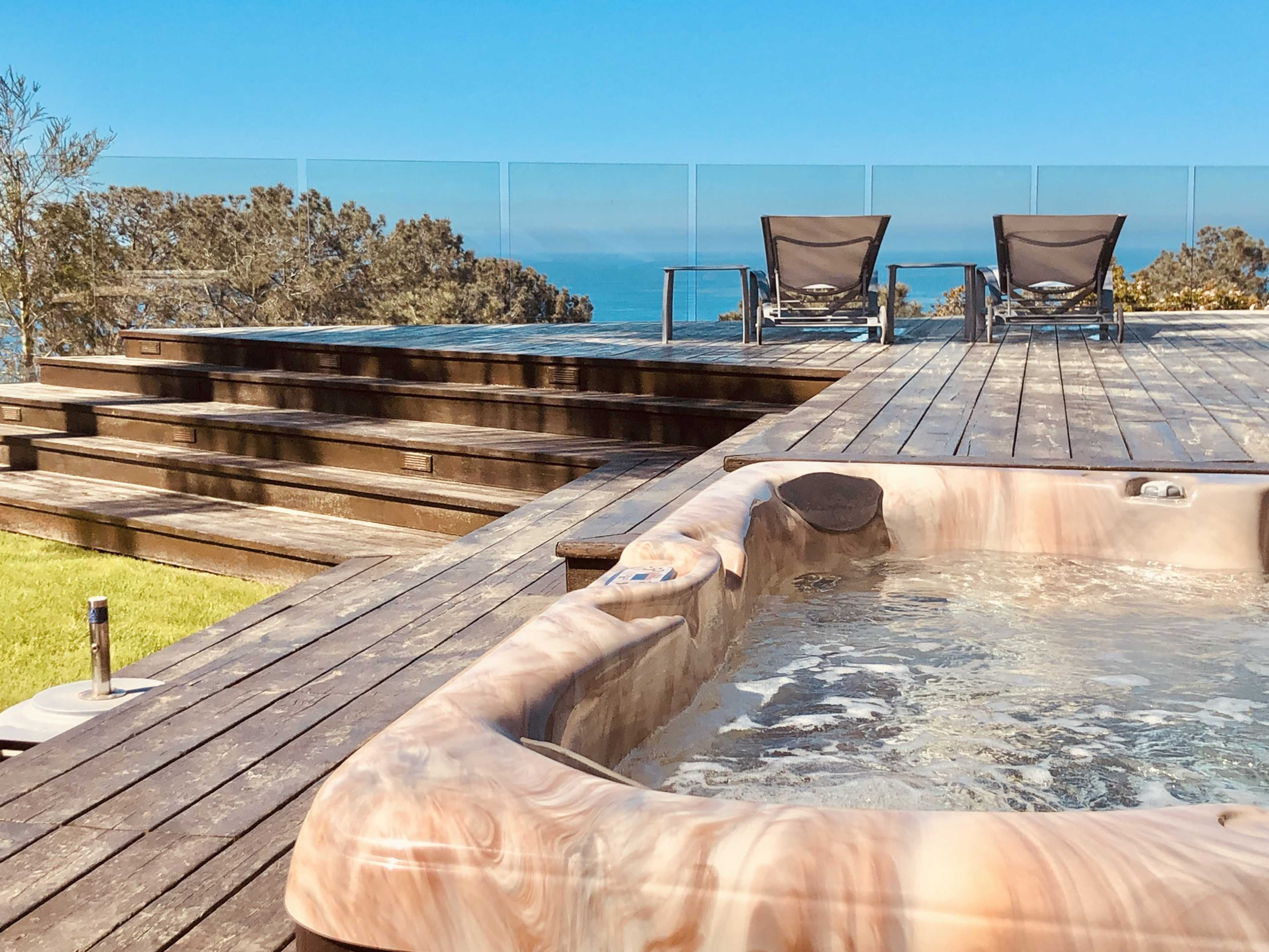 A wooden deck with a hot tub in the foreground and two lounge chairs overlooking a view of the ocean.