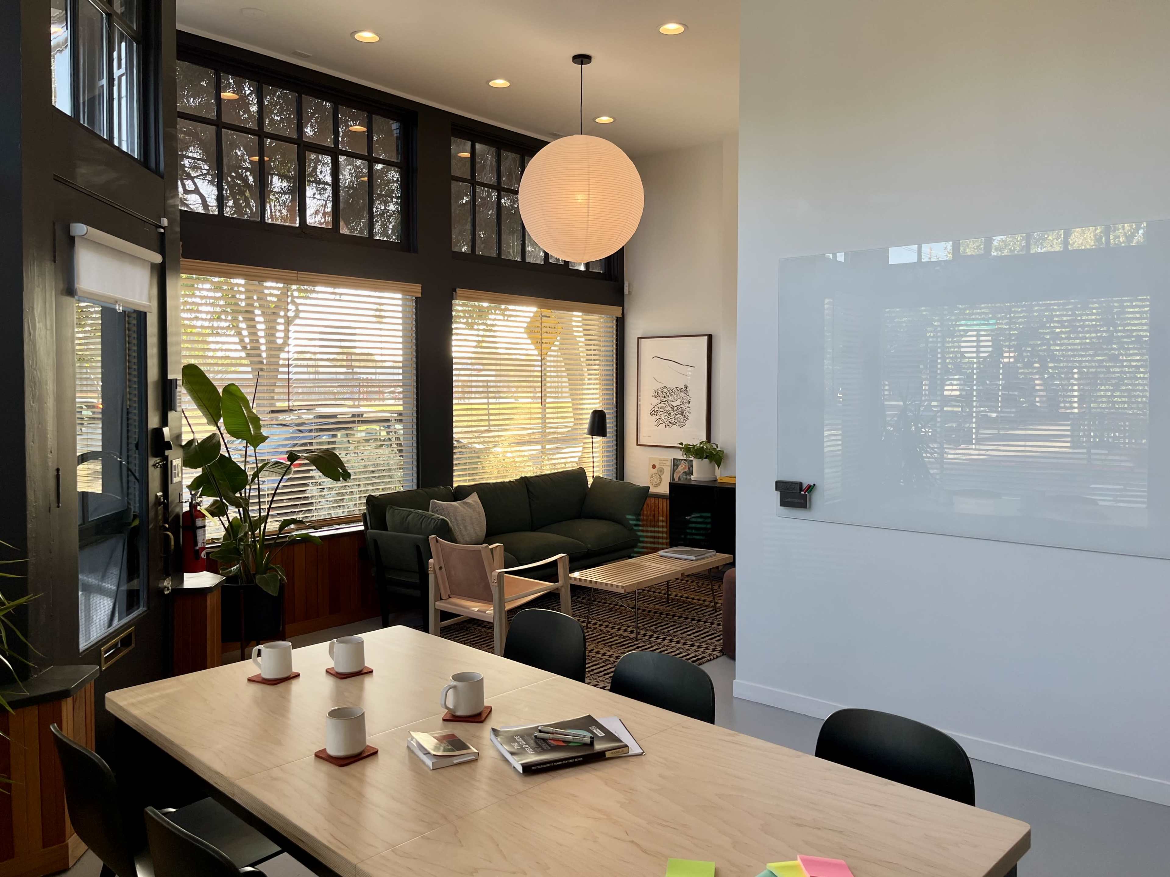A modern meeting room features a large wooden table with cups and a notepad, a seating area with a green couch, and large windows allowing natural light in.