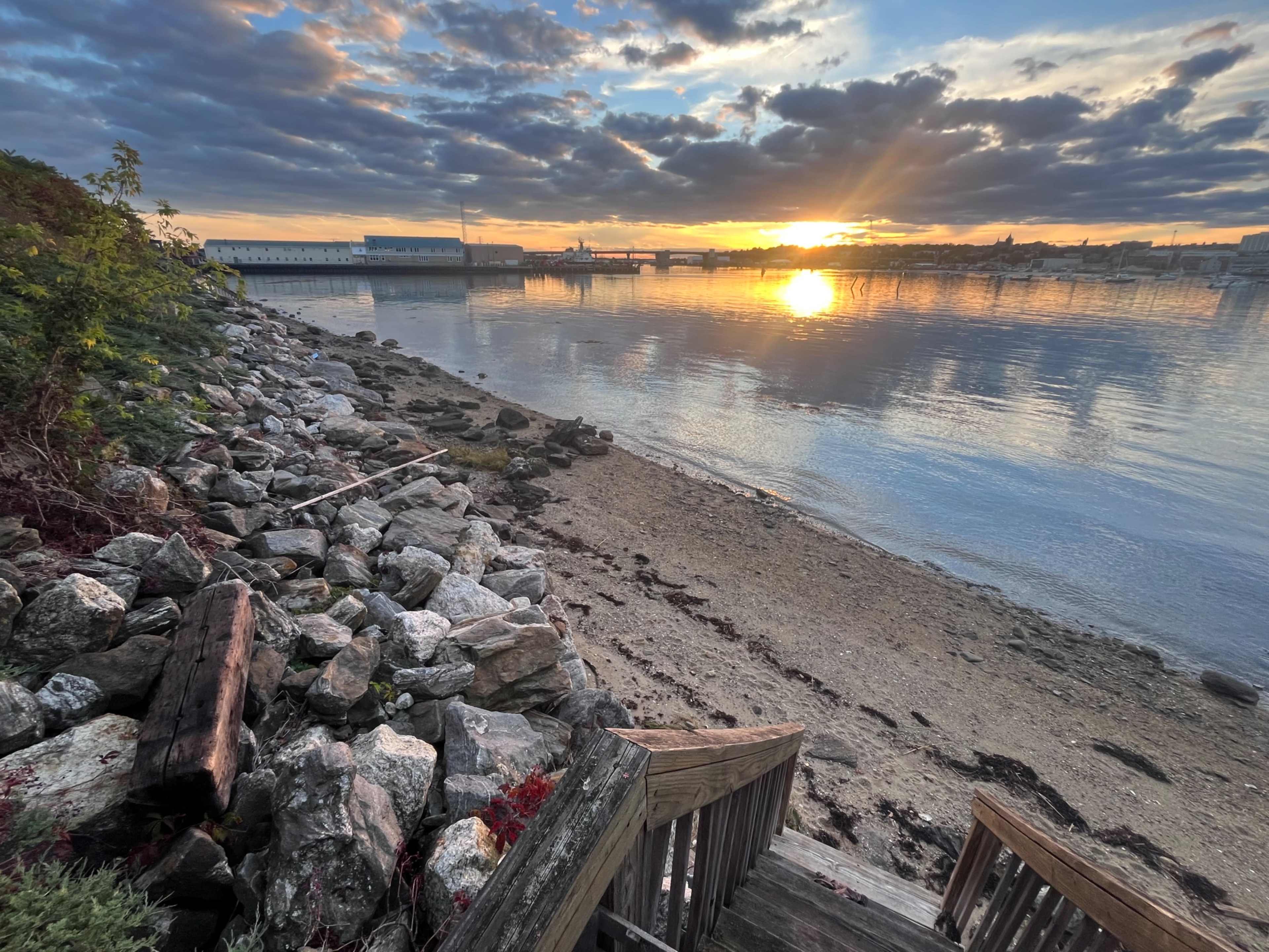 The image shows a rocky shoreline at sunset, with calm waters reflecting the colorful sky and a wooden staircase leading down to the beach.