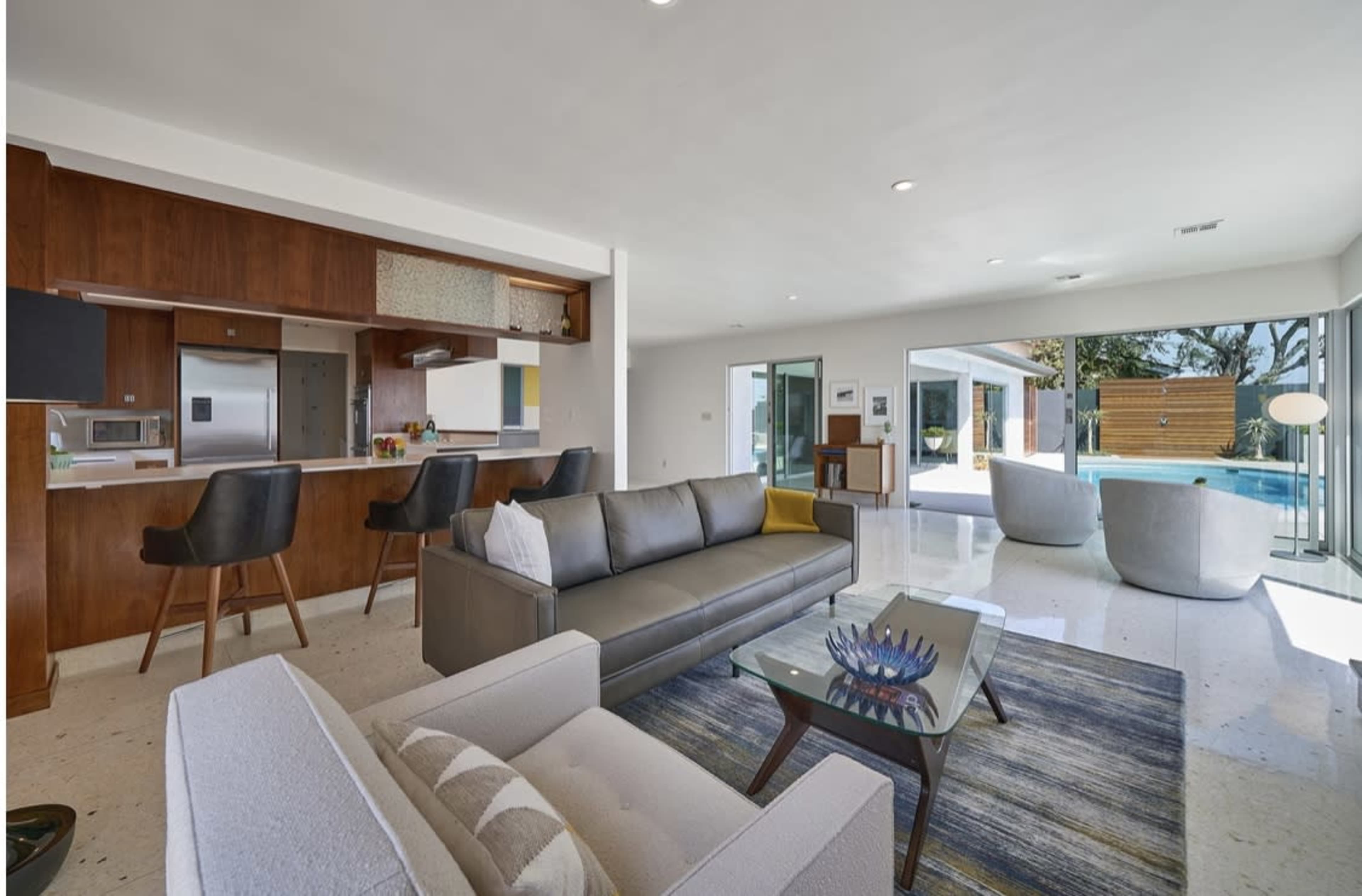 A modern living room with a gray sofa, an armchair, a glass coffee table, and an open kitchen area featuring dark wood cabinetry and bar stools.
