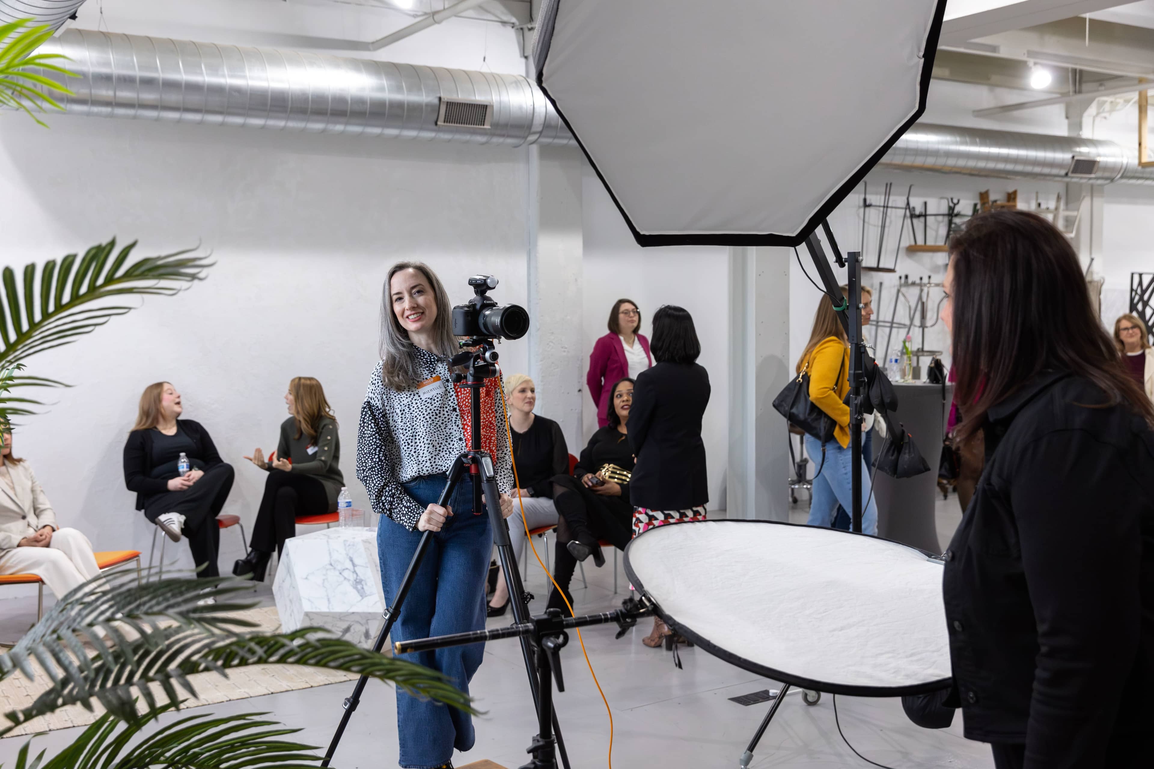 A woman smiles while holding a camera in a photo studio filled with people seated and engaging in conversation.