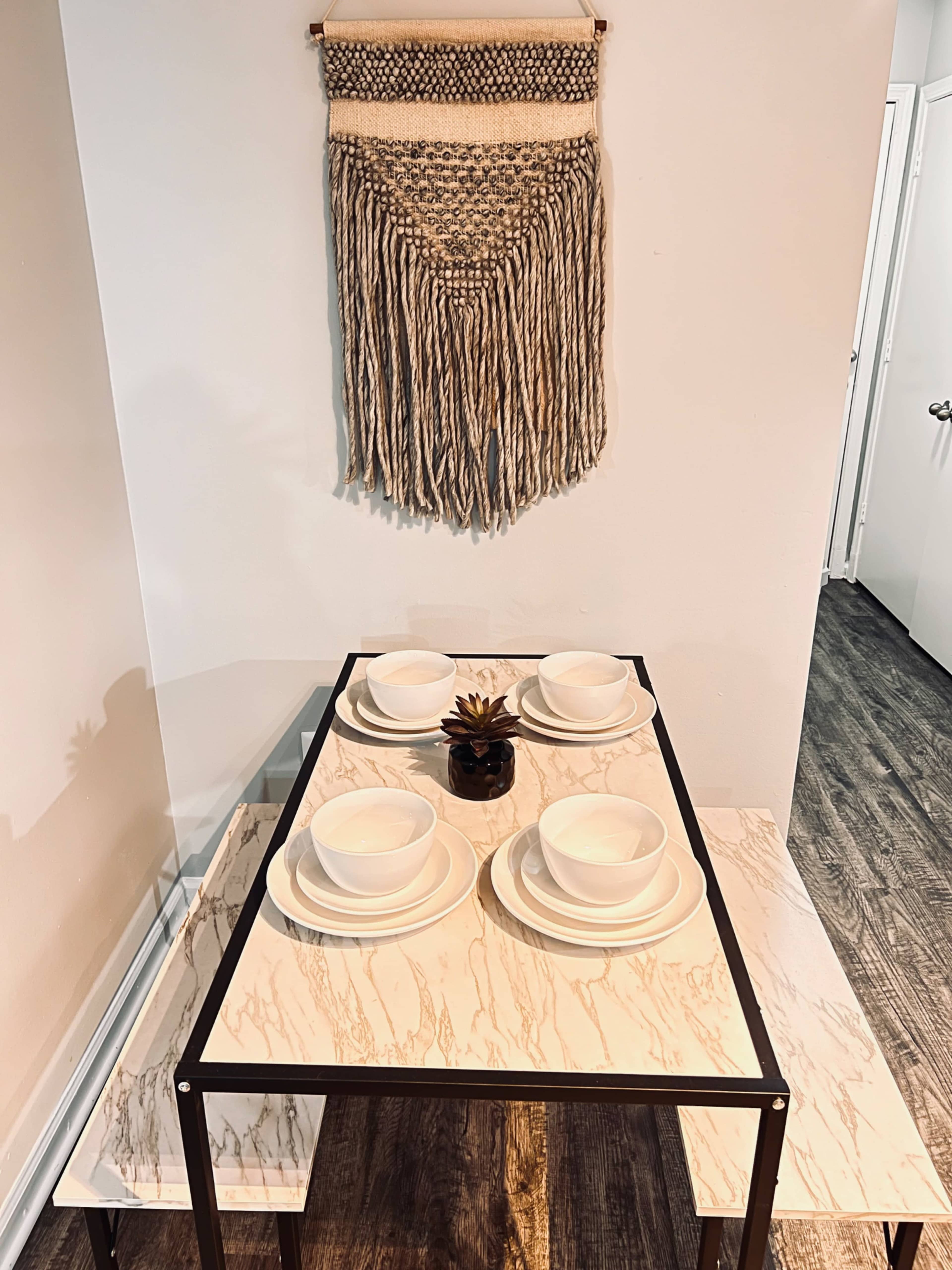 A marble-patterned dining table is set with four white plates and a small plant centerpiece, accompanied by a woven wall hanging above.