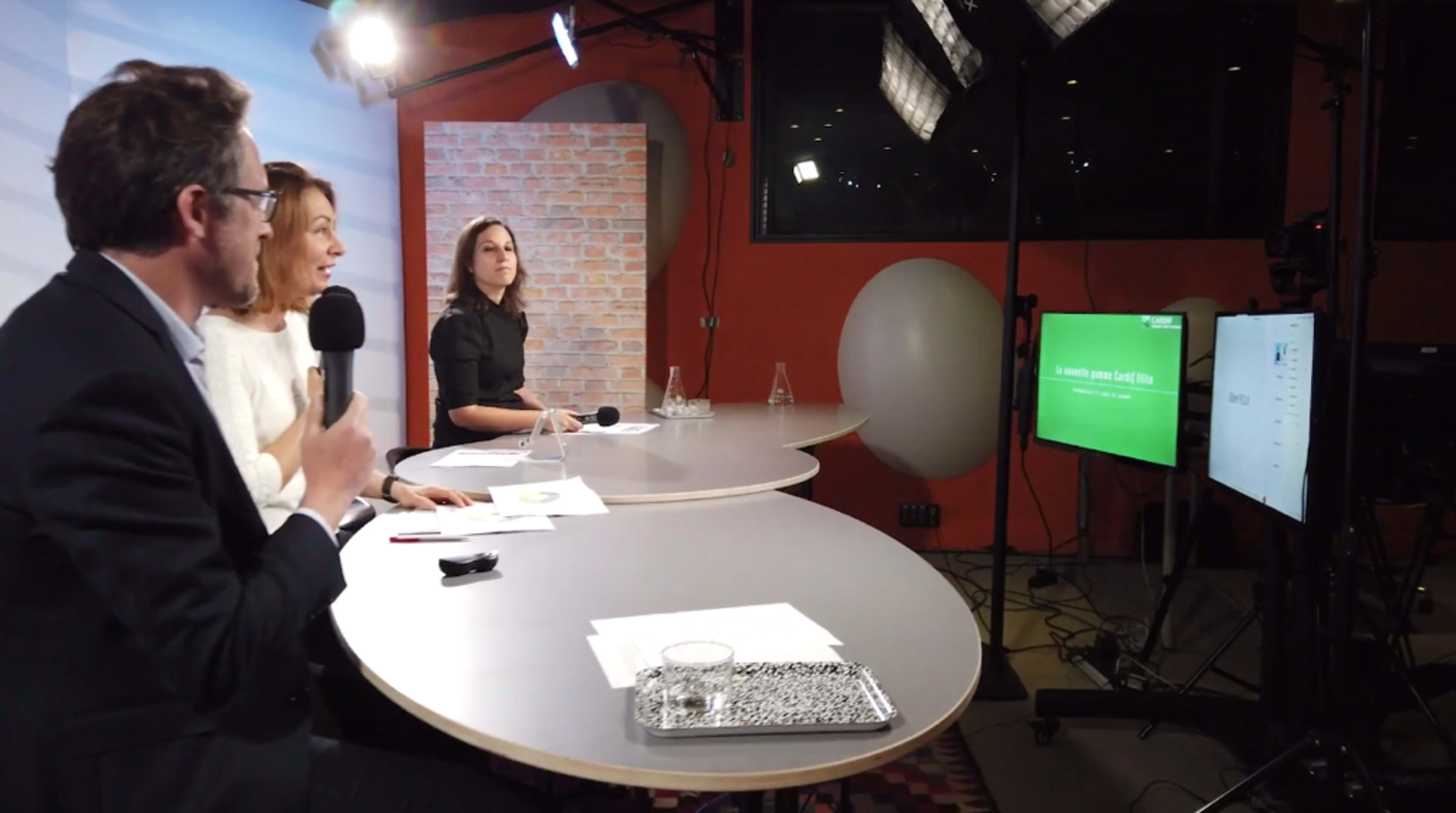 Three people sit at a table during a panel discussion, with a green screen display visible in the background.