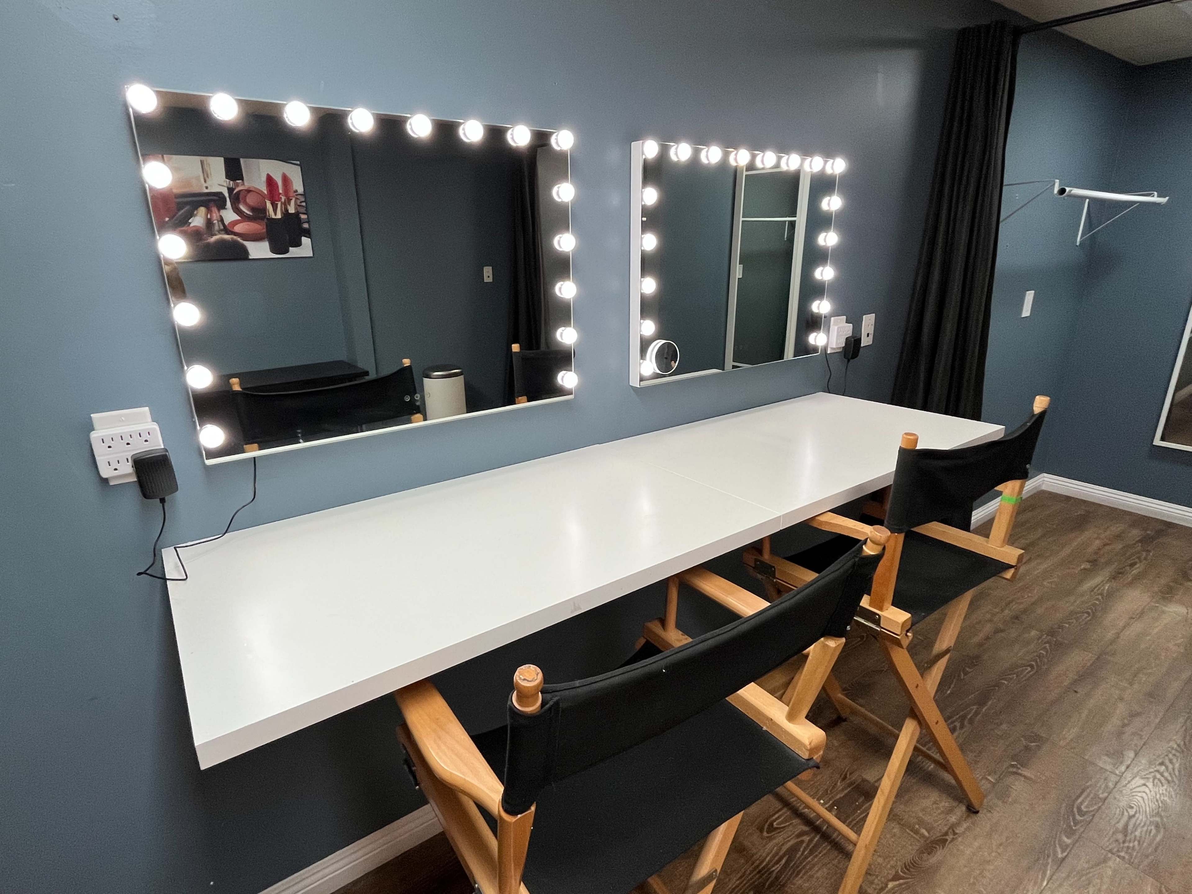 A well-lit makeup station with two mirrors and two director's chairs against a blue wall.