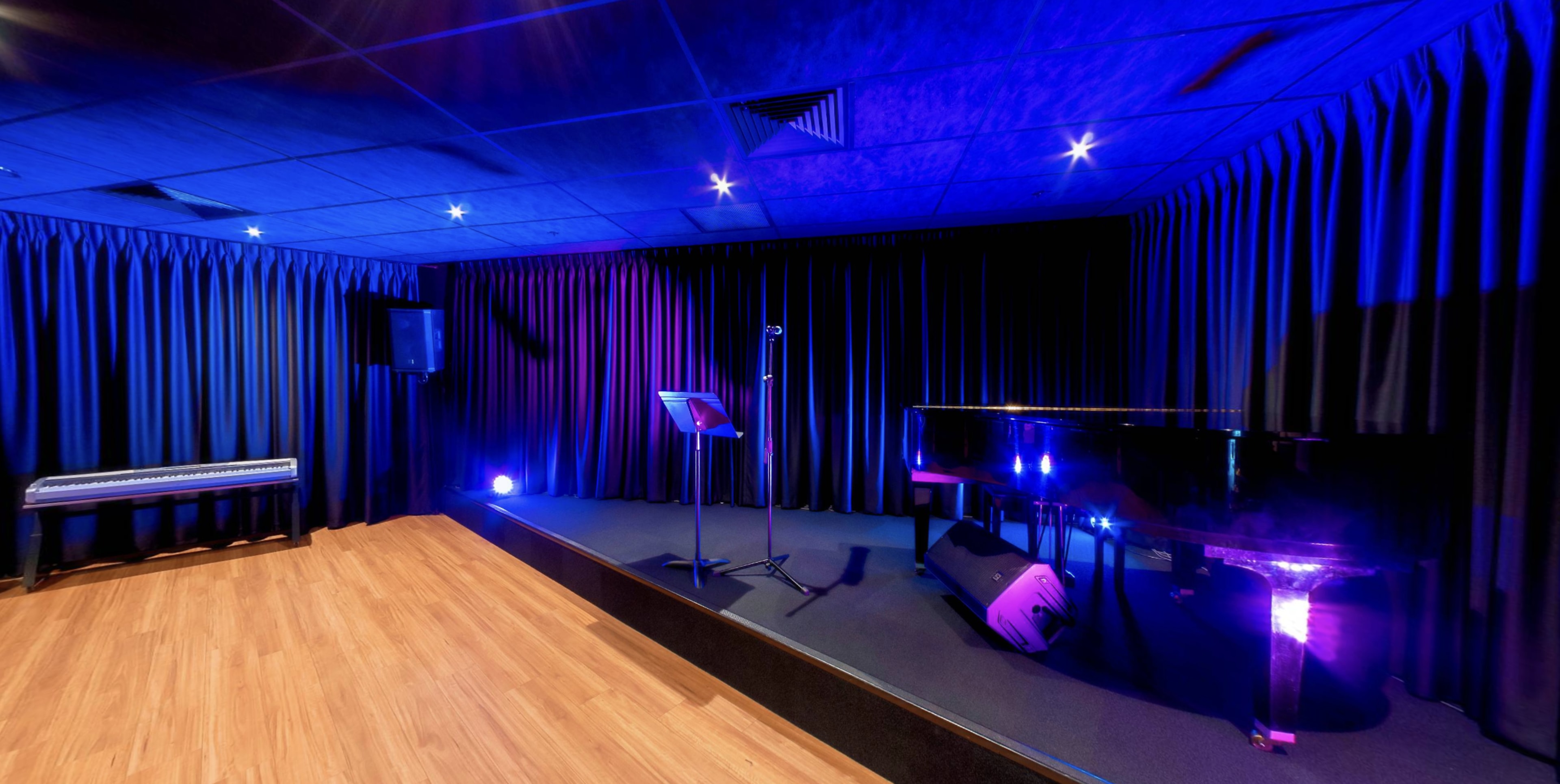 A dimly lit rehearsal space features a grand piano, a microphone stand, and a keyboard, all set against black curtains and illuminated by purple stage lights.
