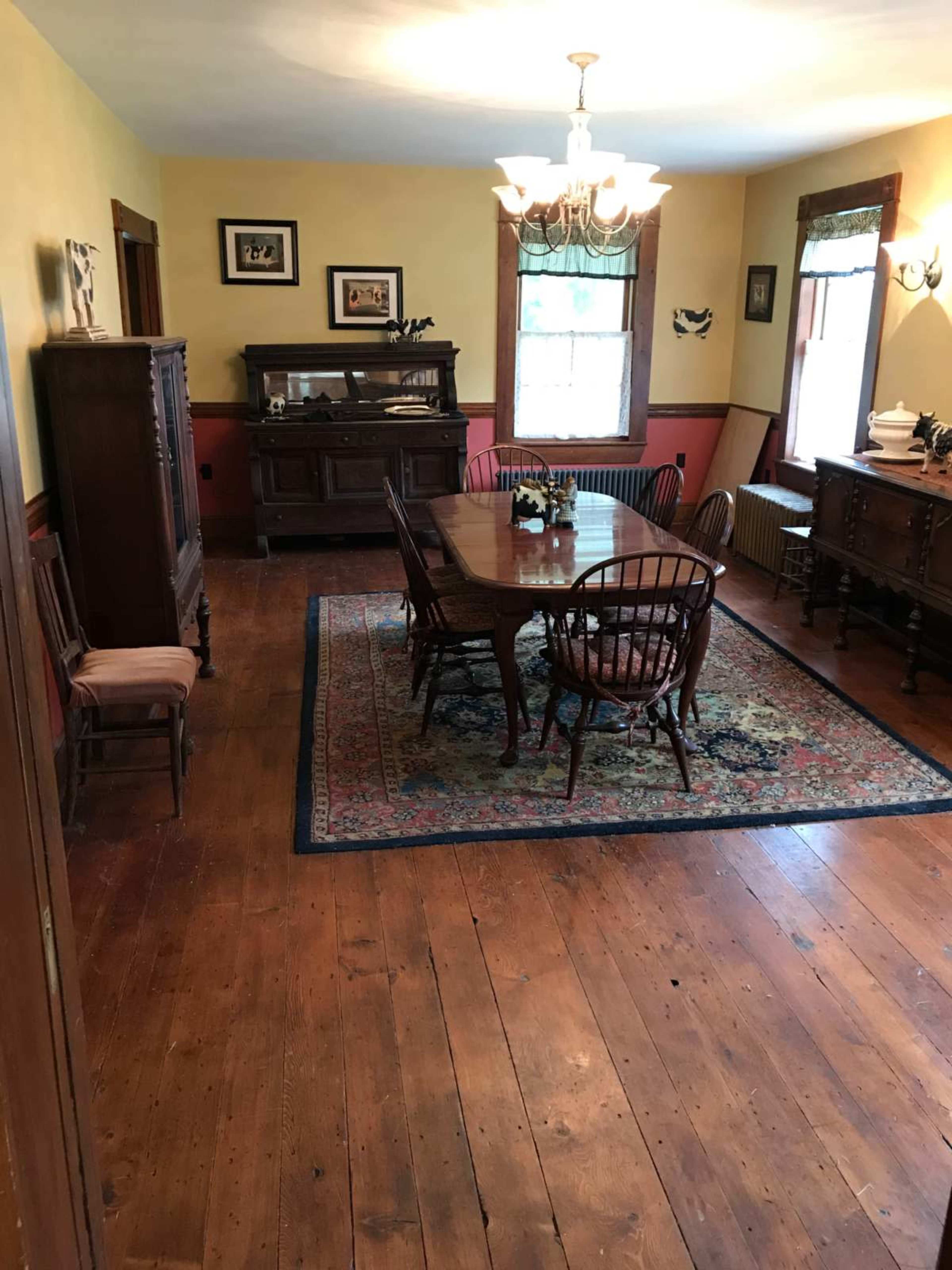 A dining room features a large wooden table surrounded by chairs, with a sideboard and display cabinet against the walls, and a colorful rug on the hardwood floor.