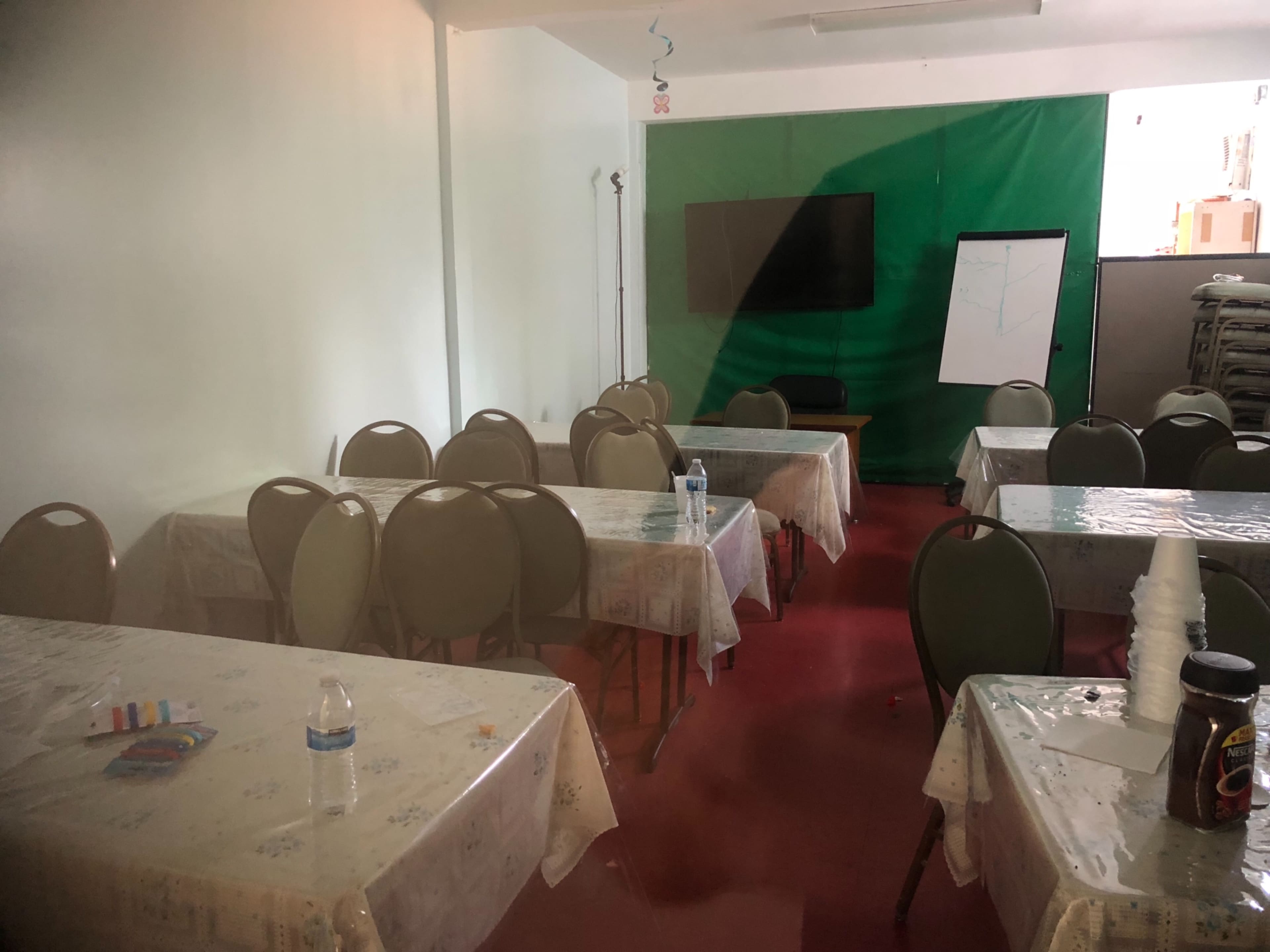 A classroom with multiple round tables covered in white tablecloths, a water bottle on one table, a whiteboard, and a large screen at the front.