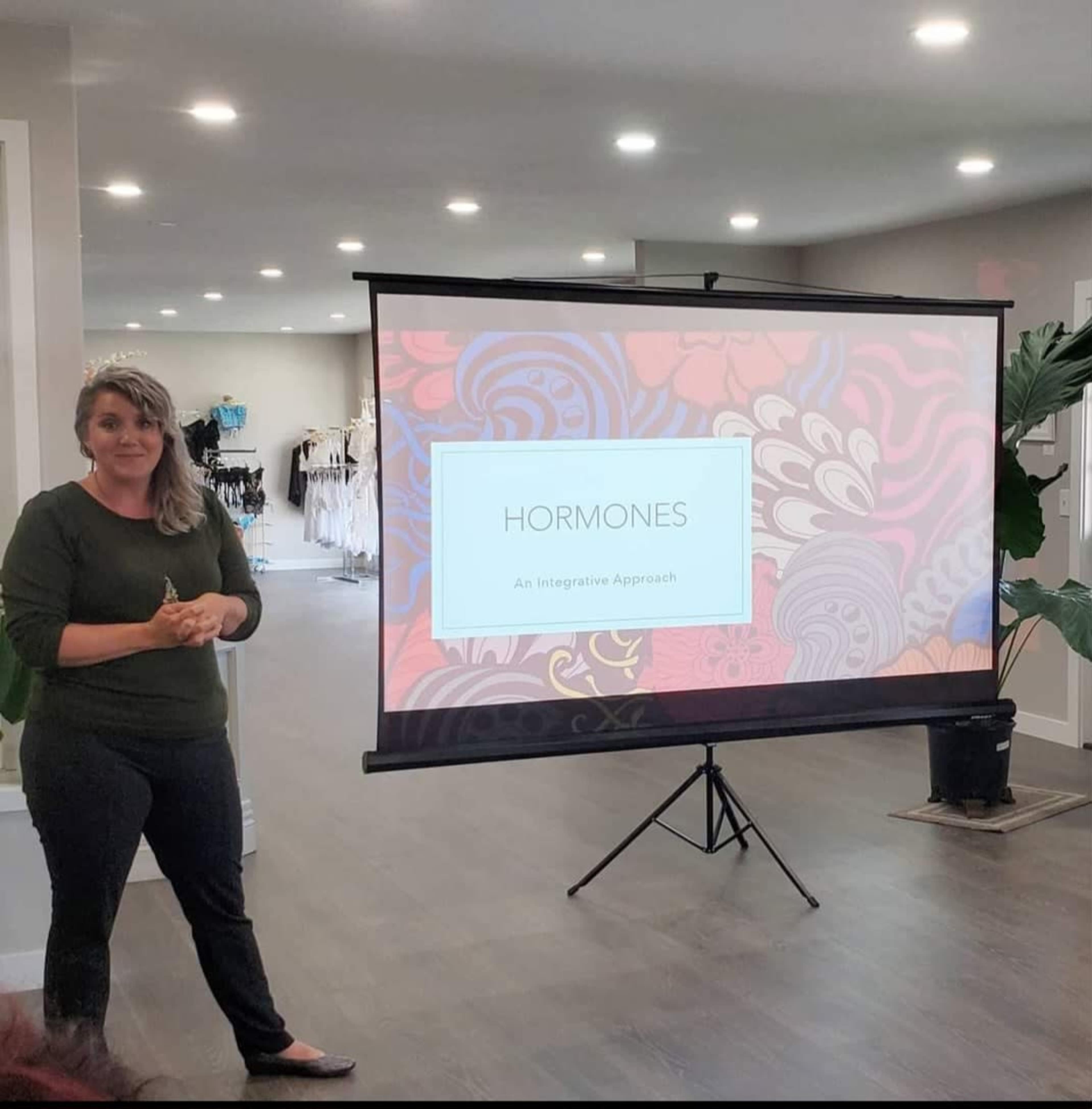 A woman stands in front of a presentation screen displaying the word "HORMONES" and the subtitle "An Integrative Approach."
