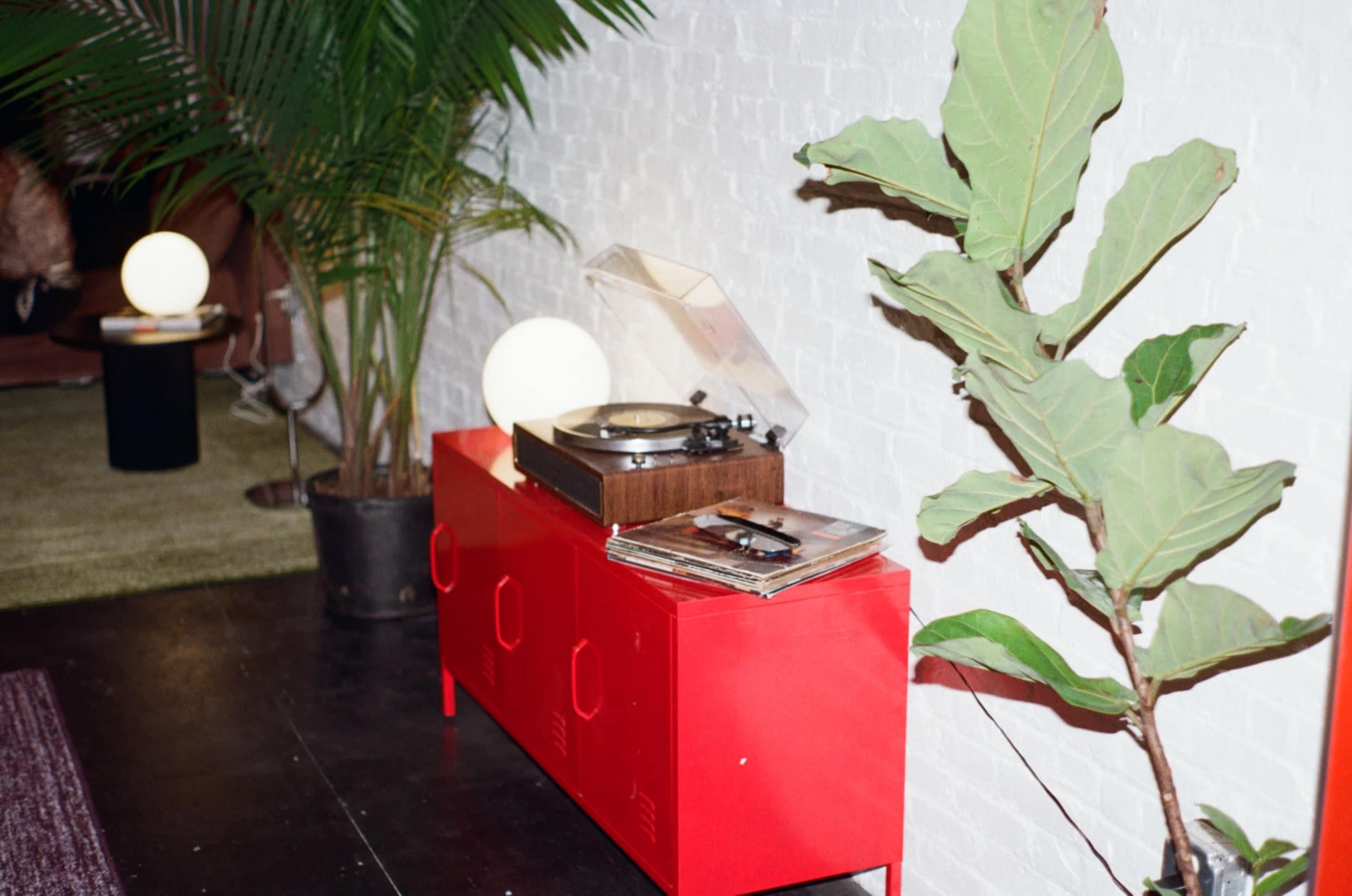 A red cabinet with a vintage record player and vinyl records sits beside a potted plant and a glowing orb lamp against a white brick wall.