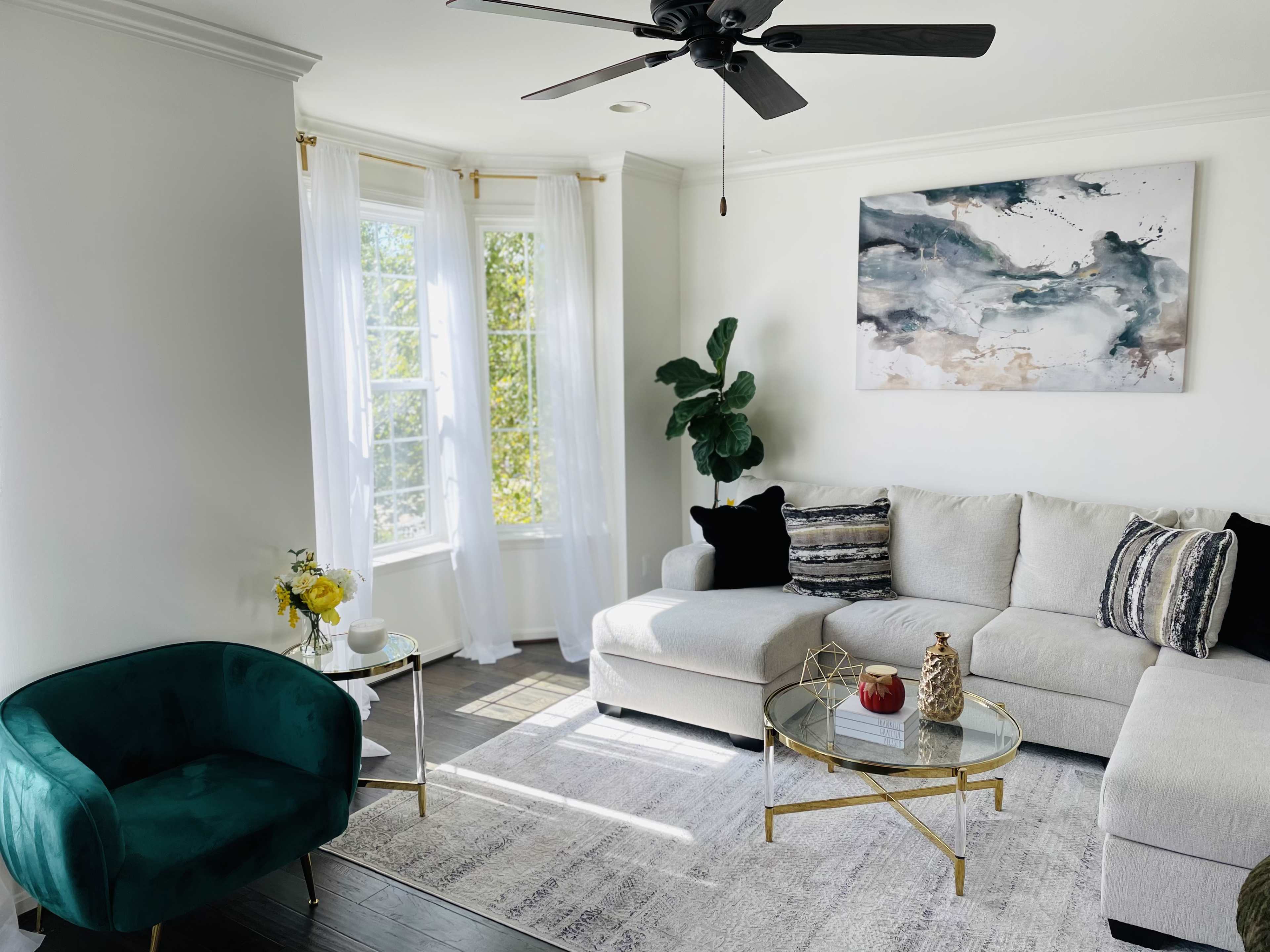 A bright living room features a white sectional sofa, a dark green armchair, a glass coffee table, and a large window with sheer curtains, complemented by a colorful wall art piece.