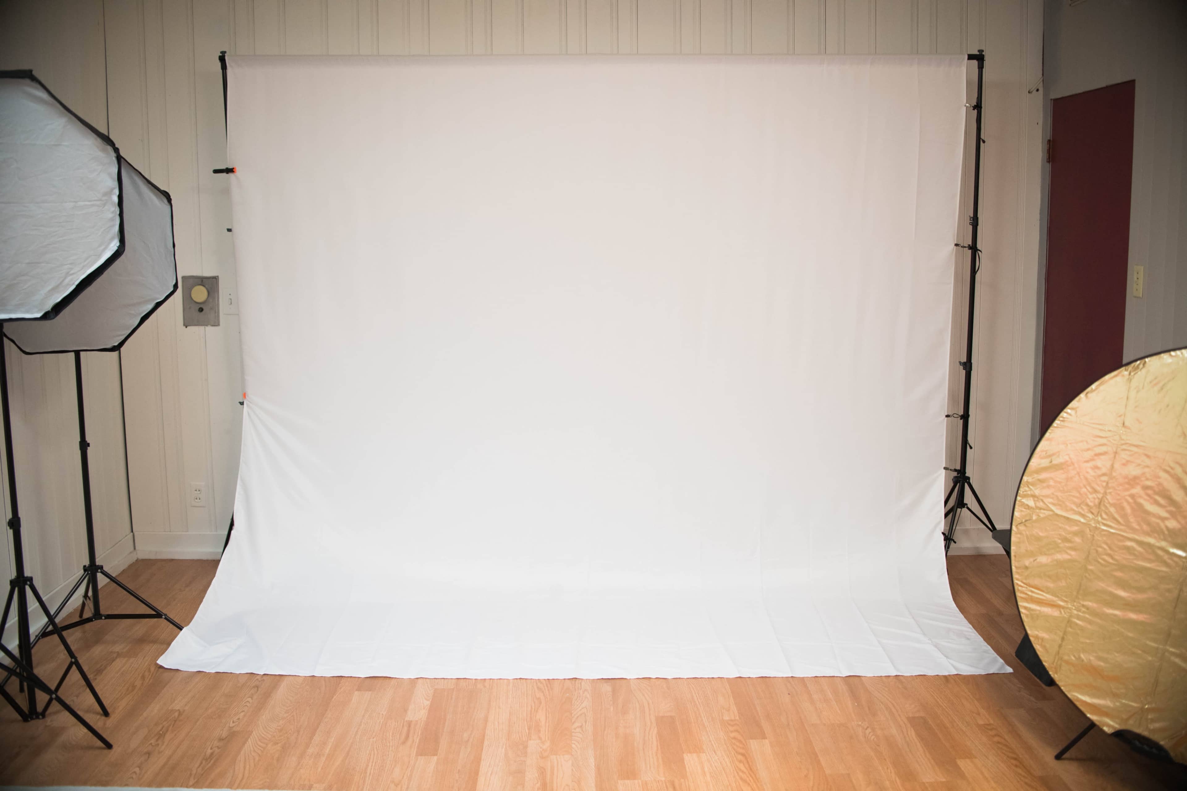 The image shows a photography studio with a large white backdrop and various lighting equipment positioned around the space.