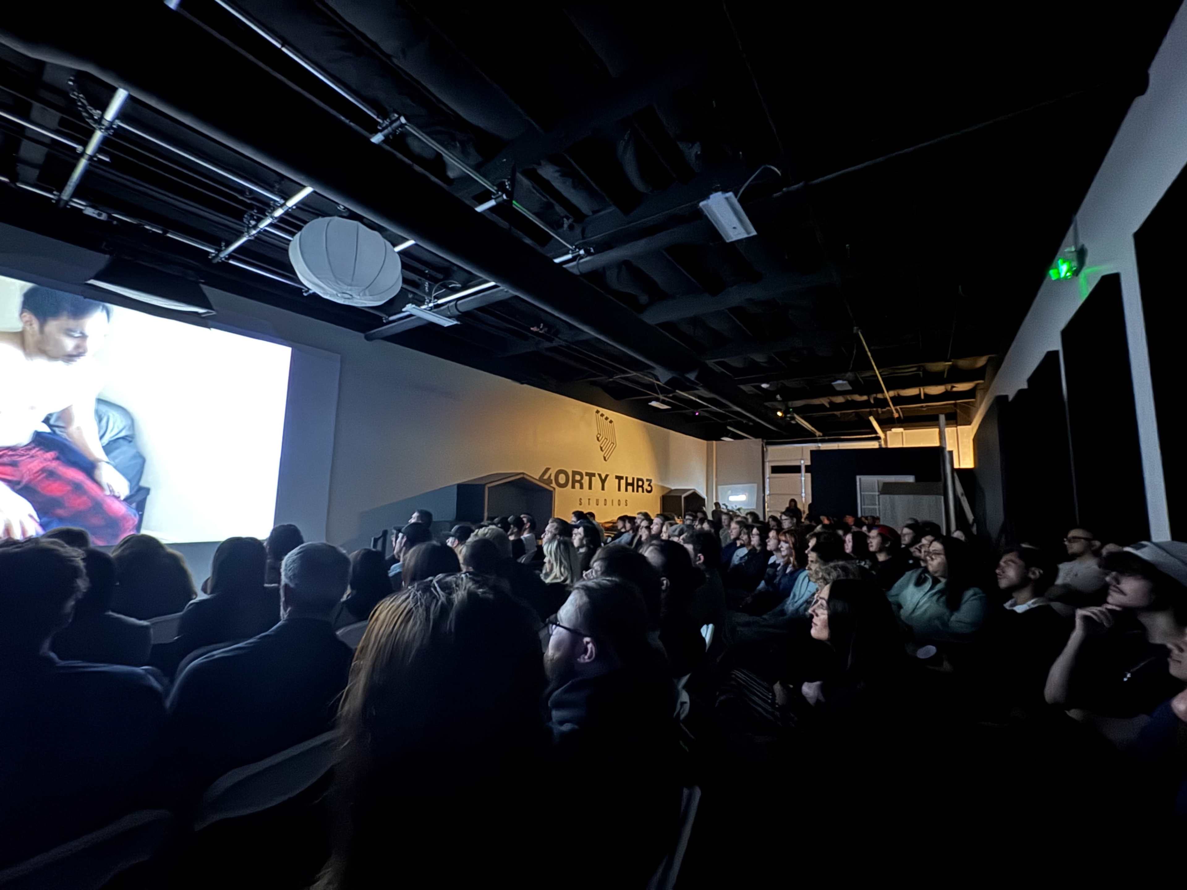 A large audience watches a presentation on a screen in a dimly lit, modern event space.