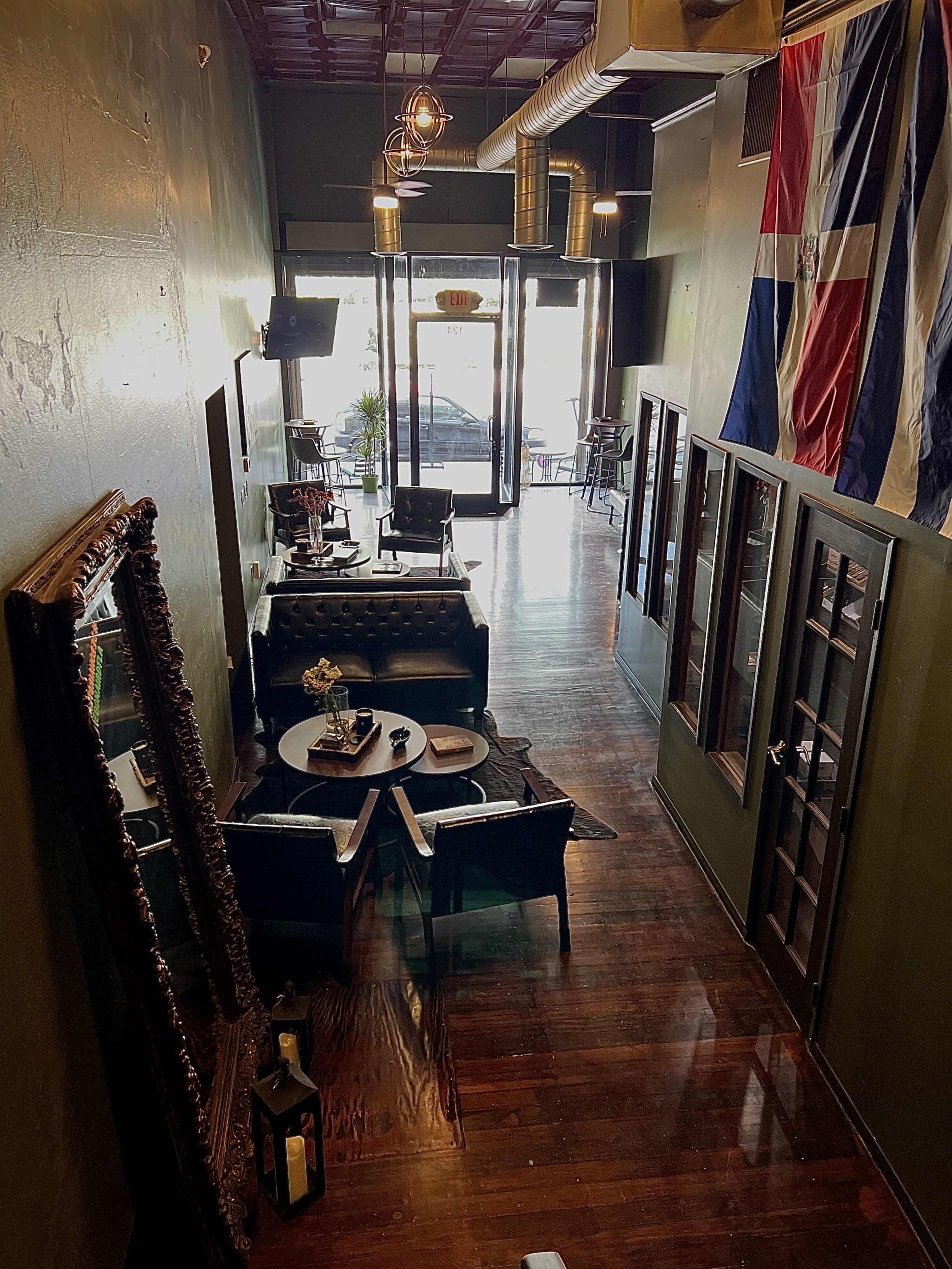 A cozy lounge area with dark wood flooring, leather seating, and a large decorative mirror, leading to a glass entrance with a visible flag hanging on the wall.